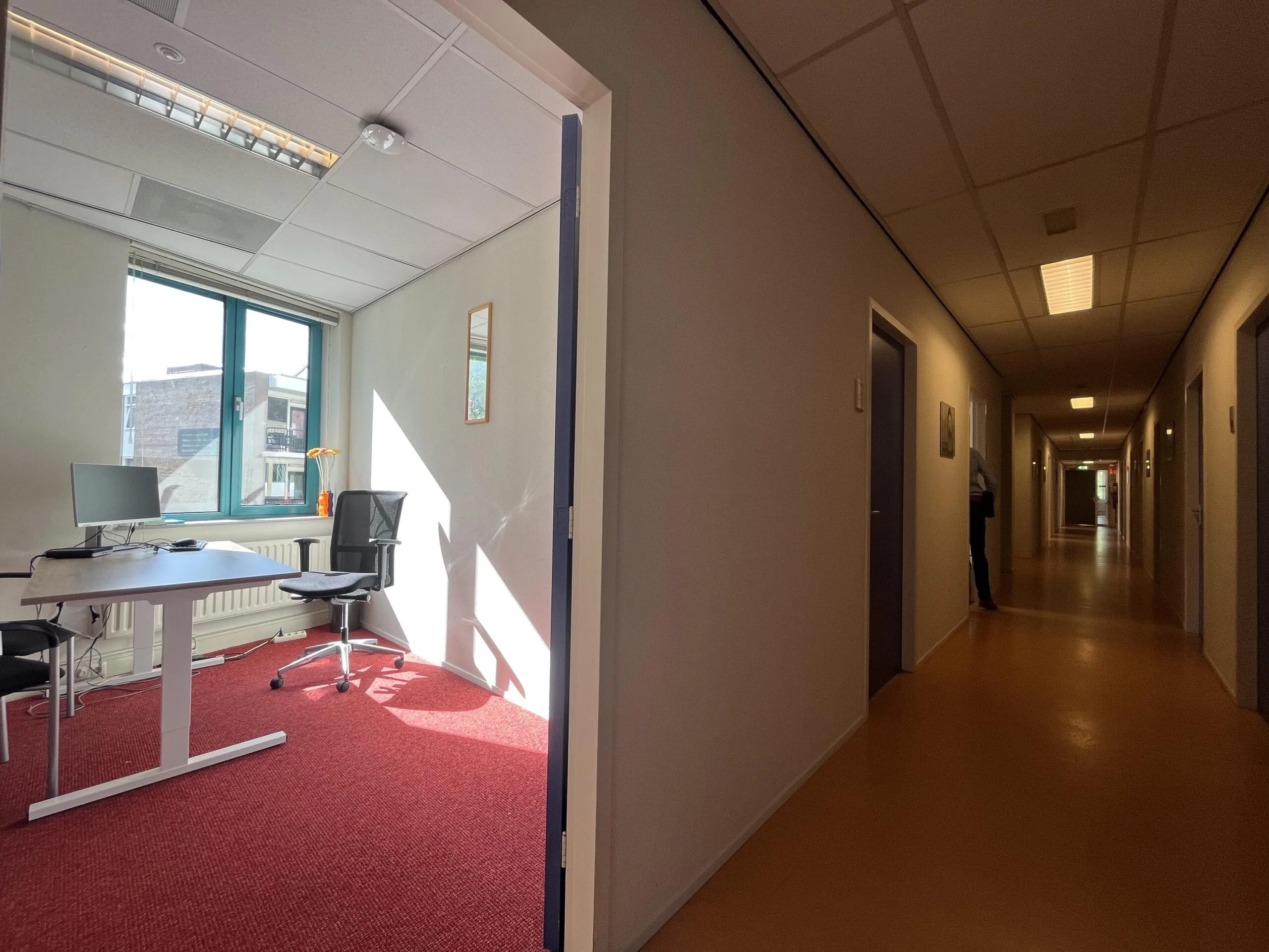 Een kantoor met een bureau en stoel kijkt uit op een lange gang met gesloten deuren.