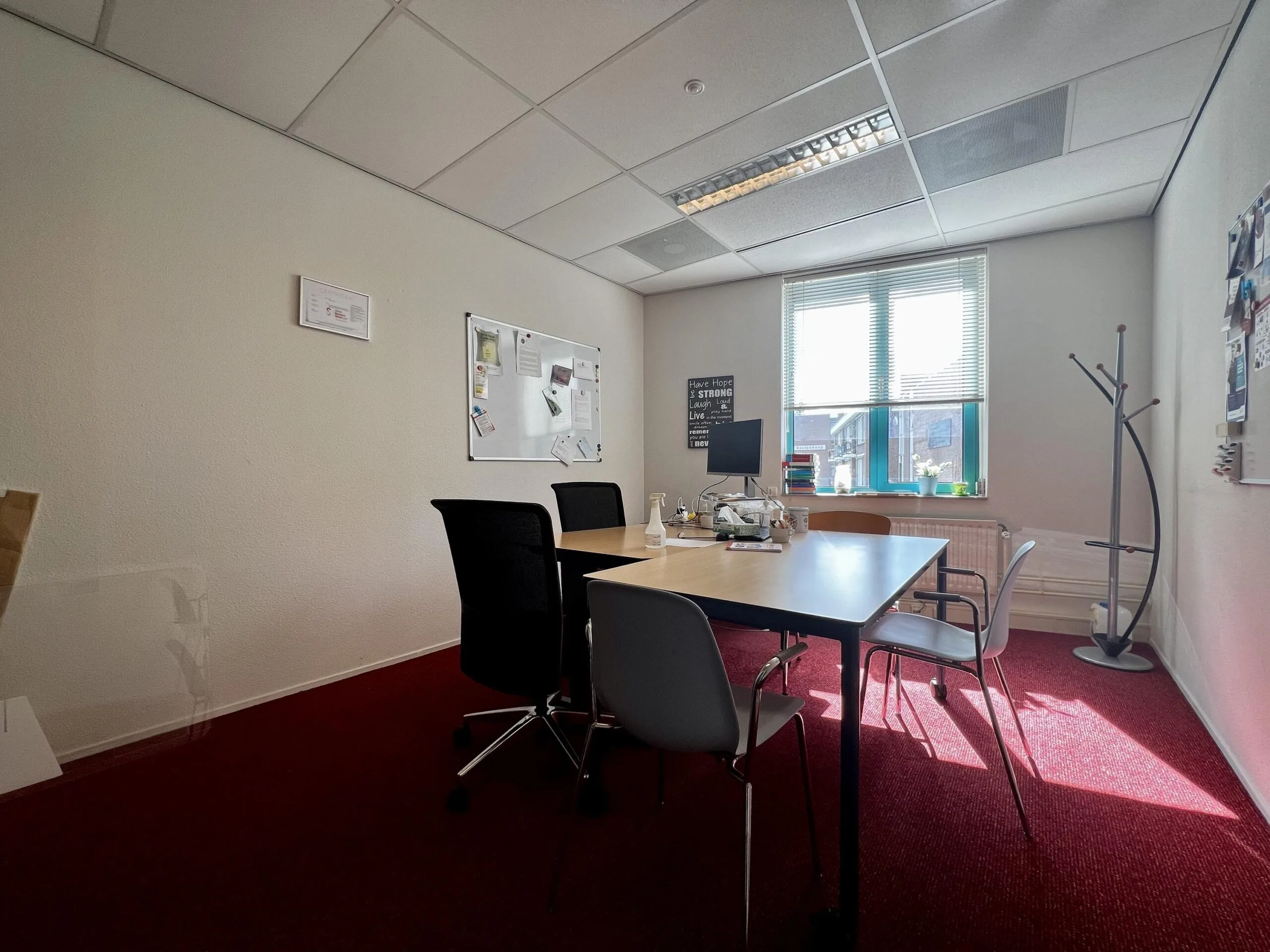 Een kleine vergaderruimte met een rood tapijt, een tafel met stoelen, en een raam dat zonlicht binnenlaat.