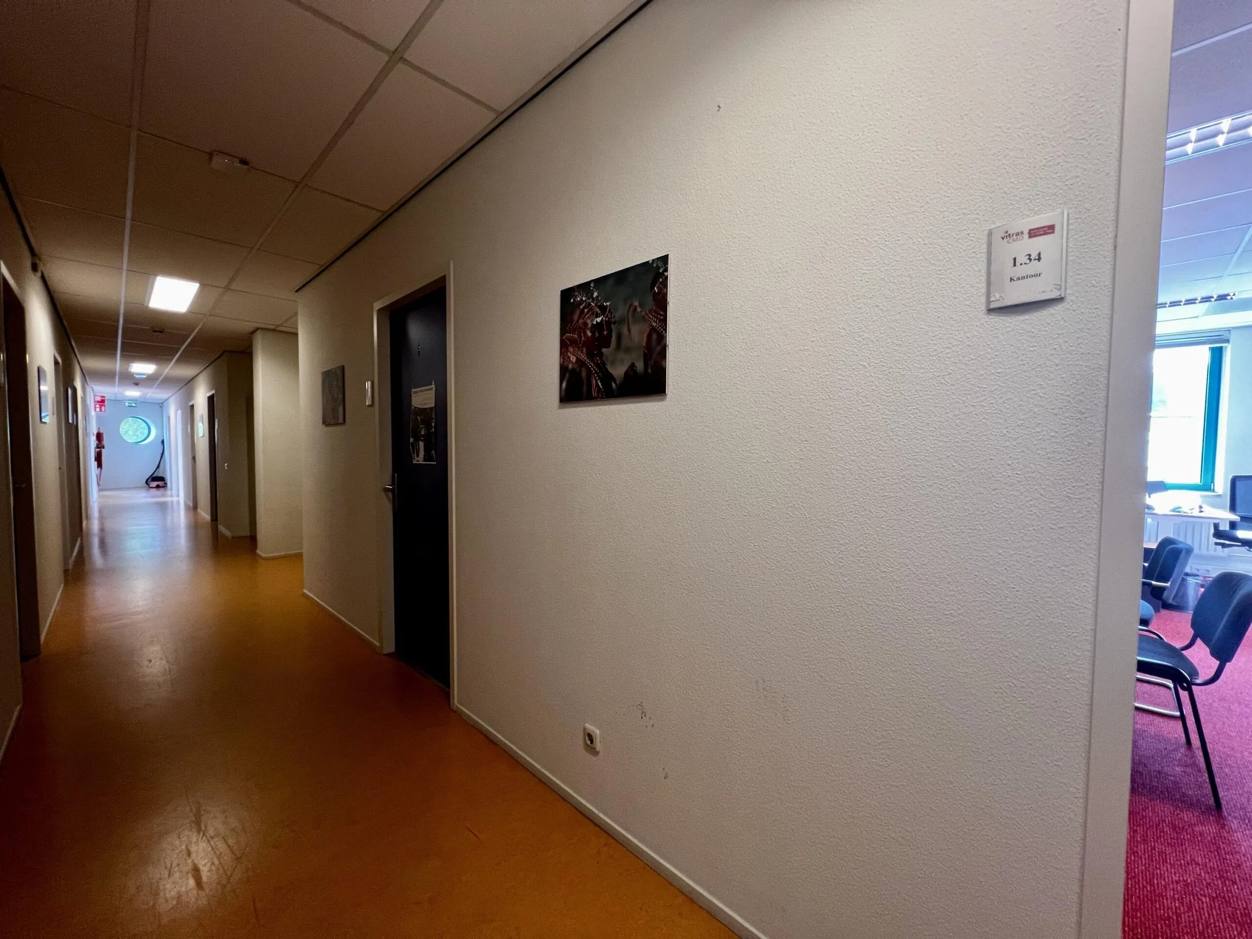 Een lange, smalle gang in een kantoor met links een brandblusser bij de nooduitgang en rechts een open deur naar een kantoorruimte.