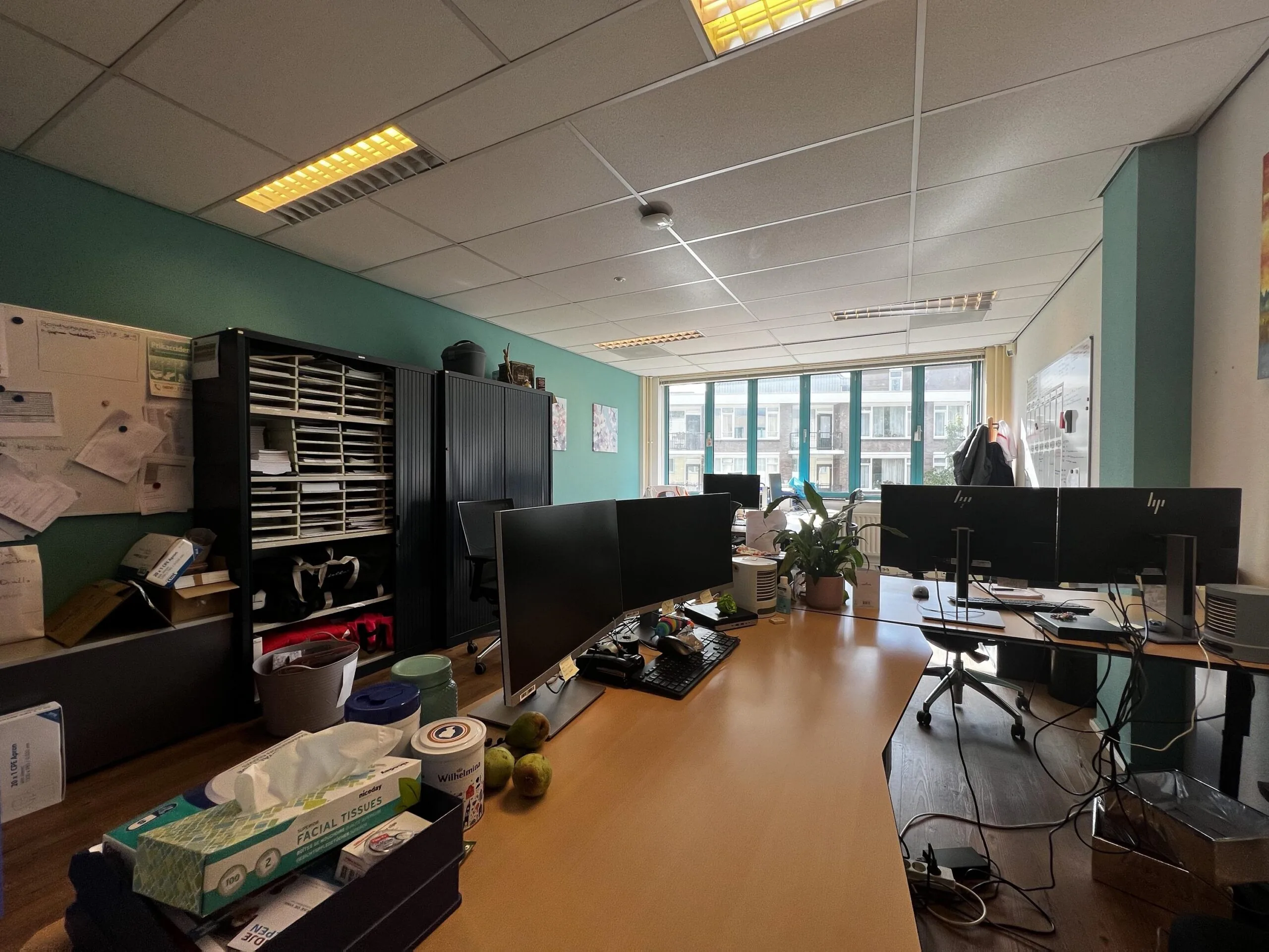 Een kantoorruimte met bureaus, computerschermen en kantoormaterialen, verlicht door grote ramen aan de achterkant.