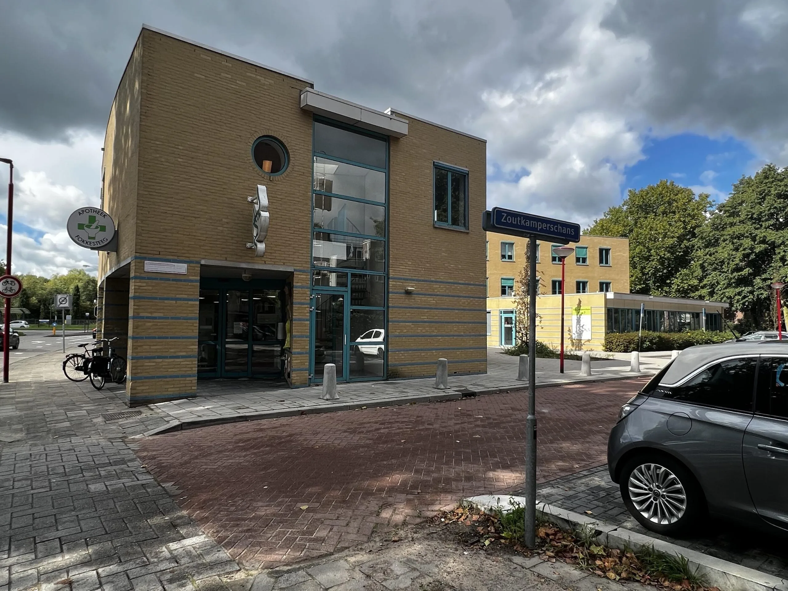 Een modern gebouw met het bord "Zoutkamperschans" en de apotheek "Vokesteeg" naast een straat met geparkeerde auto.
