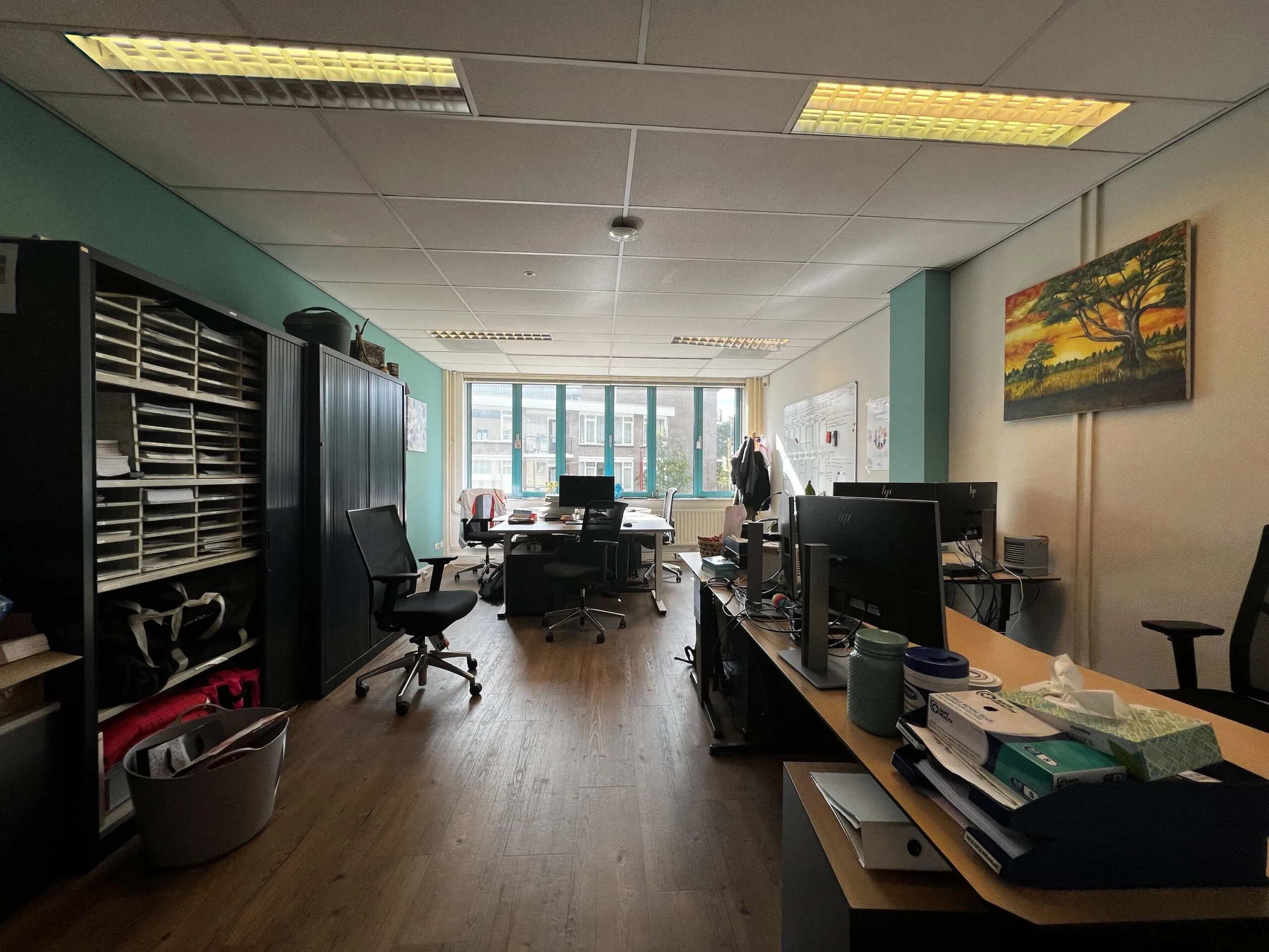 Een kantoorruimte met bureaus, stoelen, en computers, verlicht door plafondlampen, met een groot raam en schilderij aan de muur.