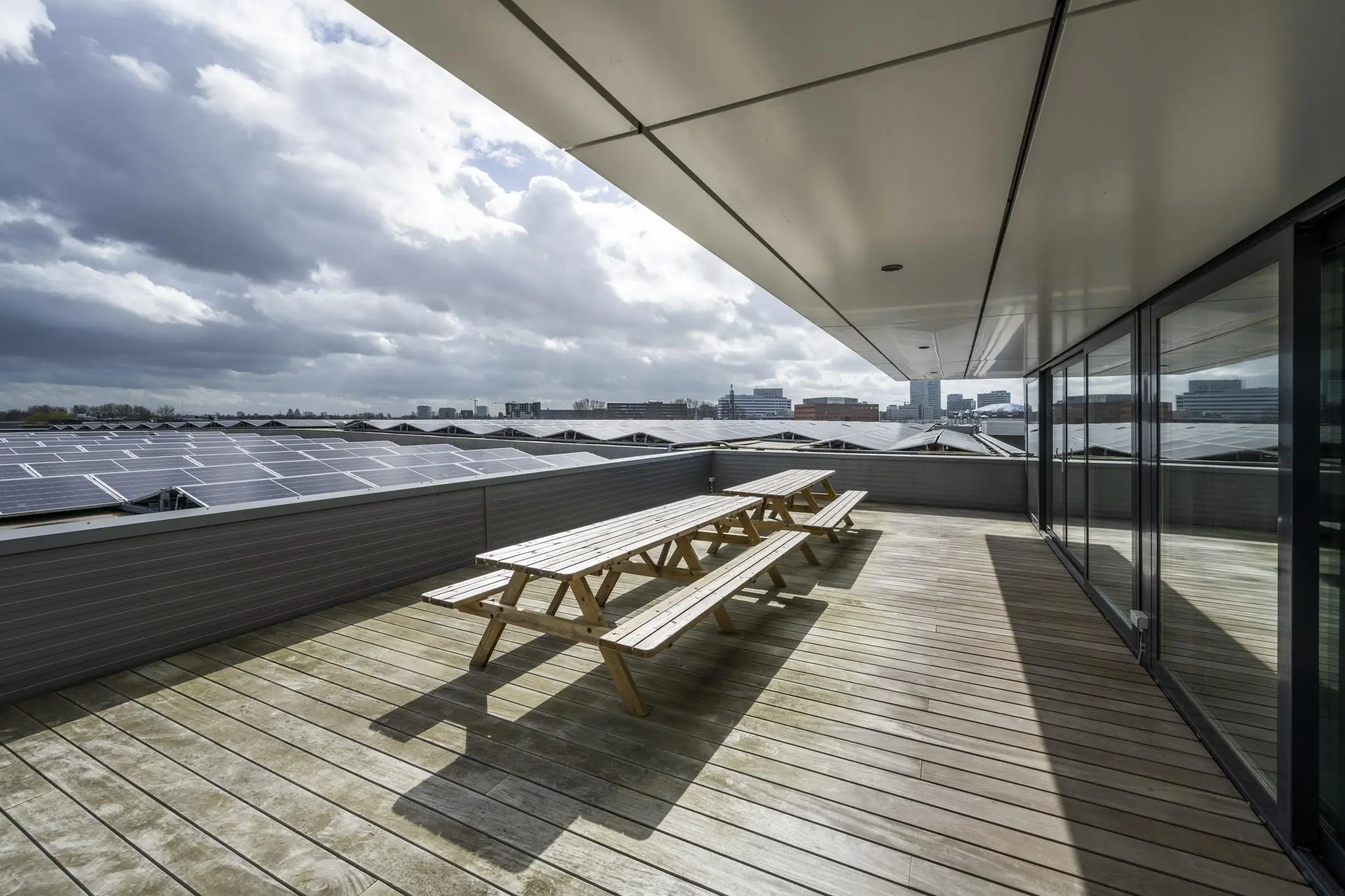 Houten dakterras met picknicktafels aan de Dynamostraat, met uitzicht op zonnepanelen en de skyline van de stad.