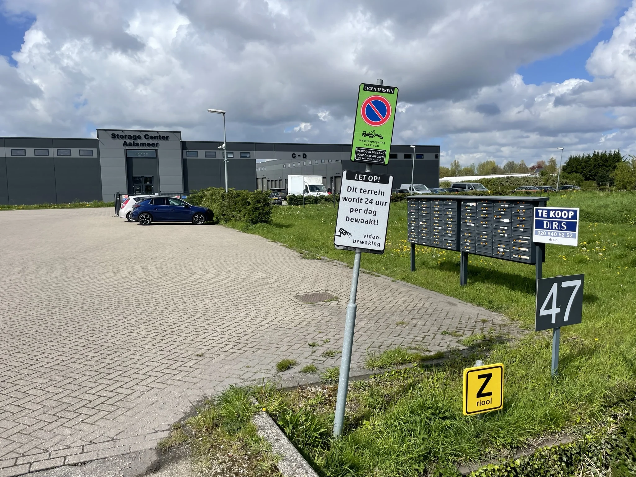 Vooraanzicht van het Storage Center Aalsmeer met parkeerplaats, brievenbussen en diverse borden, waaronder een te koop-bord en camerabewakingswaarschuwing.