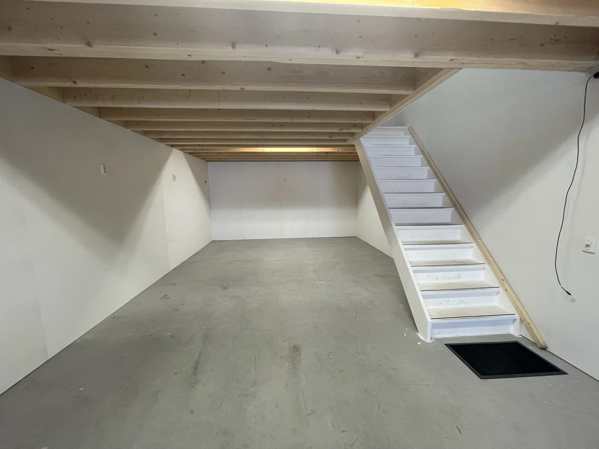 Een lege, strak afgewerkte kelderruimte met een betonnen vloer, houten plafondbalken en een witte trap aan de rechterzijde.