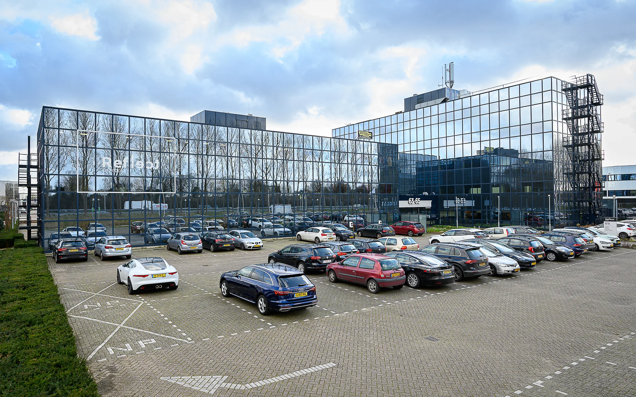 Een bedrijfsgebouw met een glazen gevel, weerspiegelend bomen, met een drukke parkeerplaats ervoor.