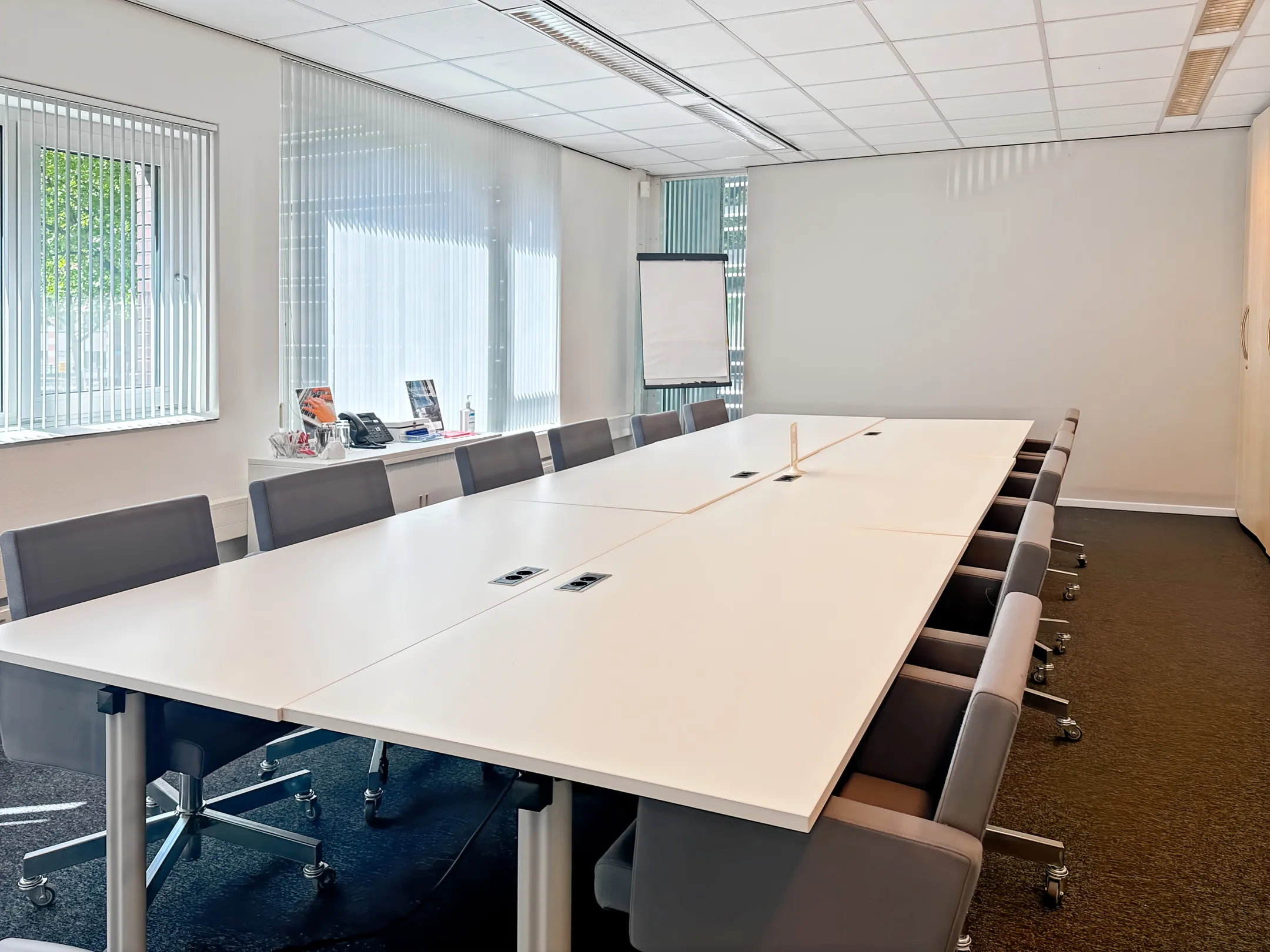 Een ruime vergaderruimte met een lange witte tafel, grijze stoelen, een flip-over en ramen met verticale jaloezieën.