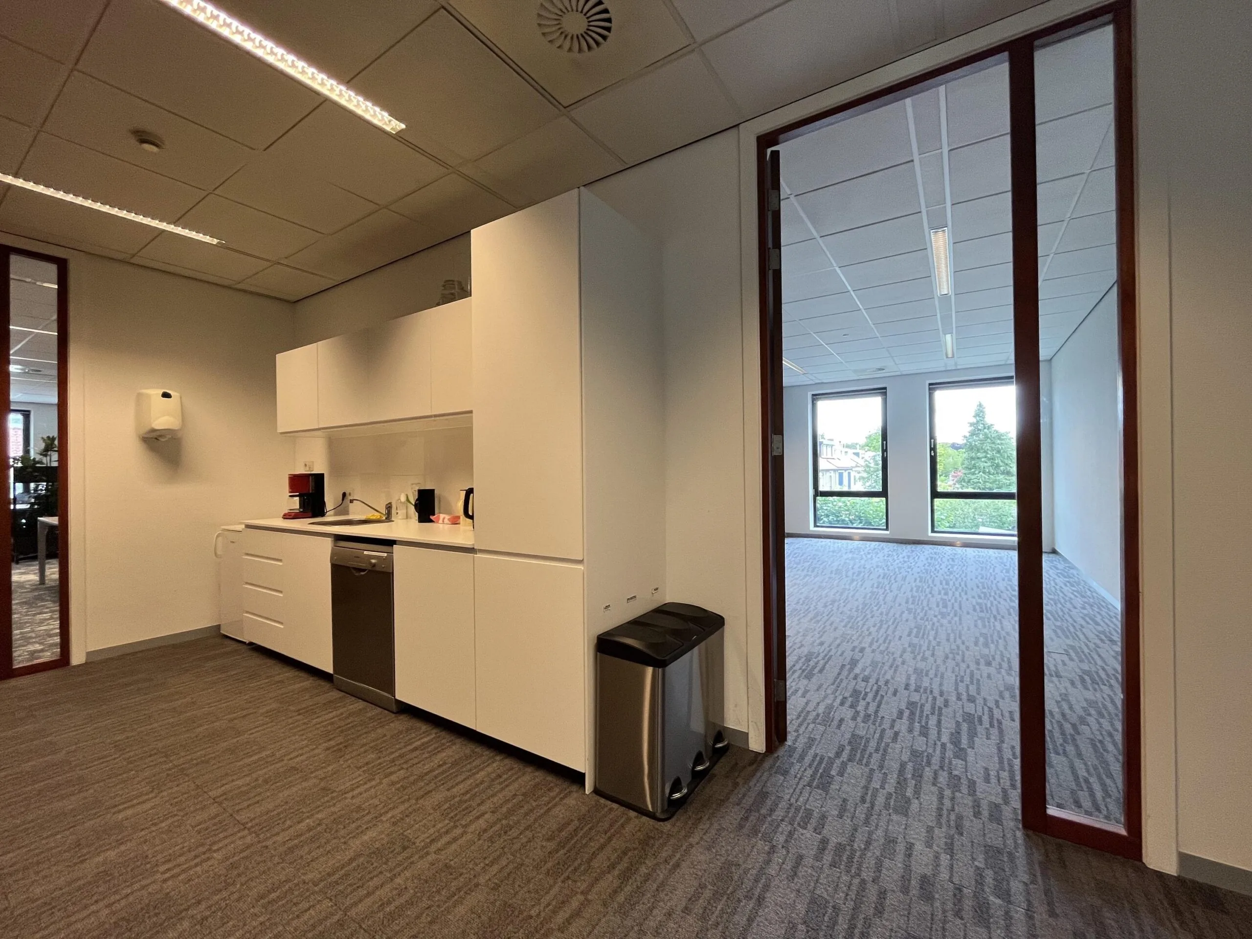 Een modern kantoor met een kleine keuken en een lege vergaderruimte met uitzicht op bomen.