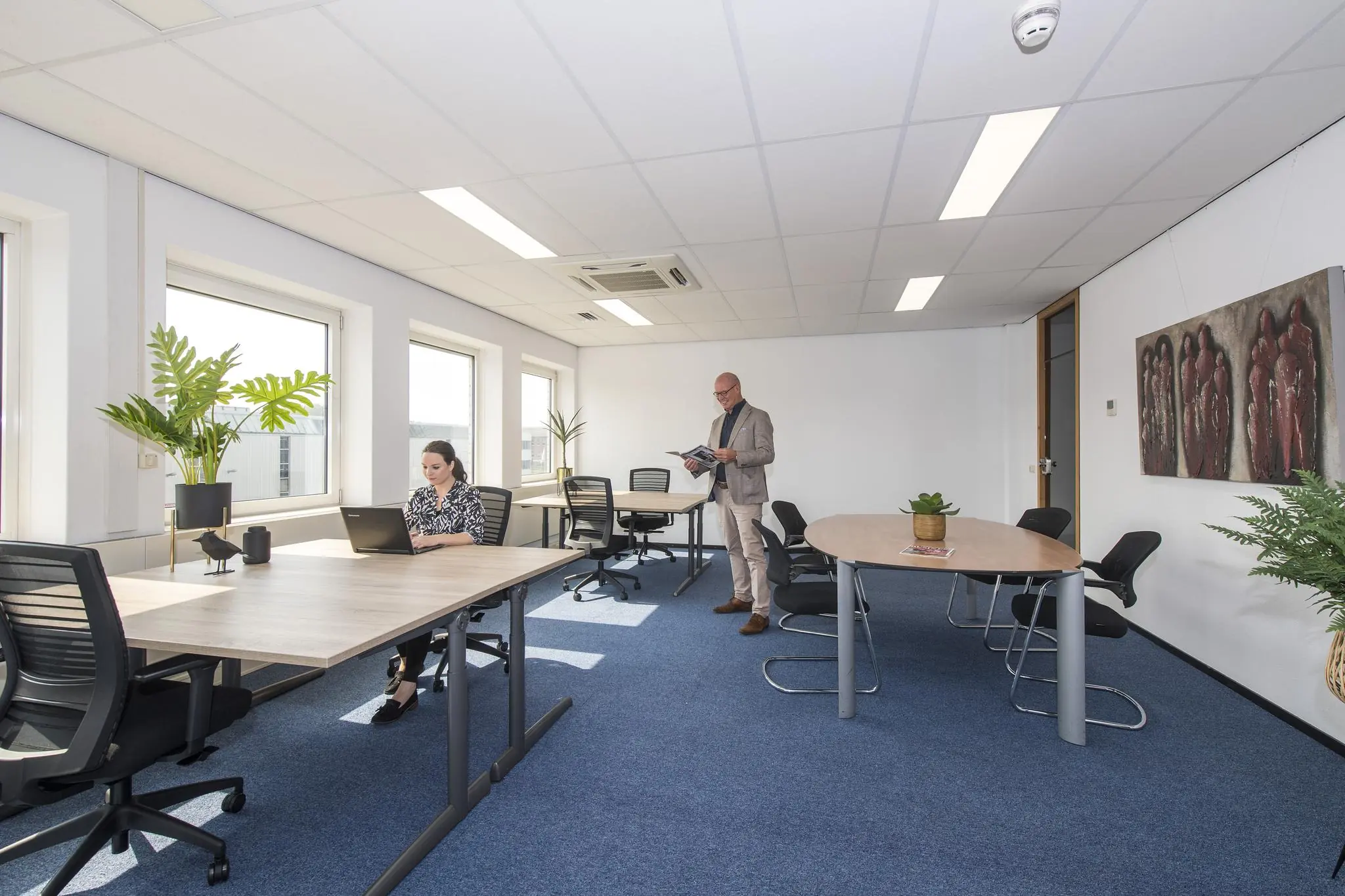 Licht en modern kantoor met een vrouw werkend aan een laptop en een man die een tijdschrift leest.