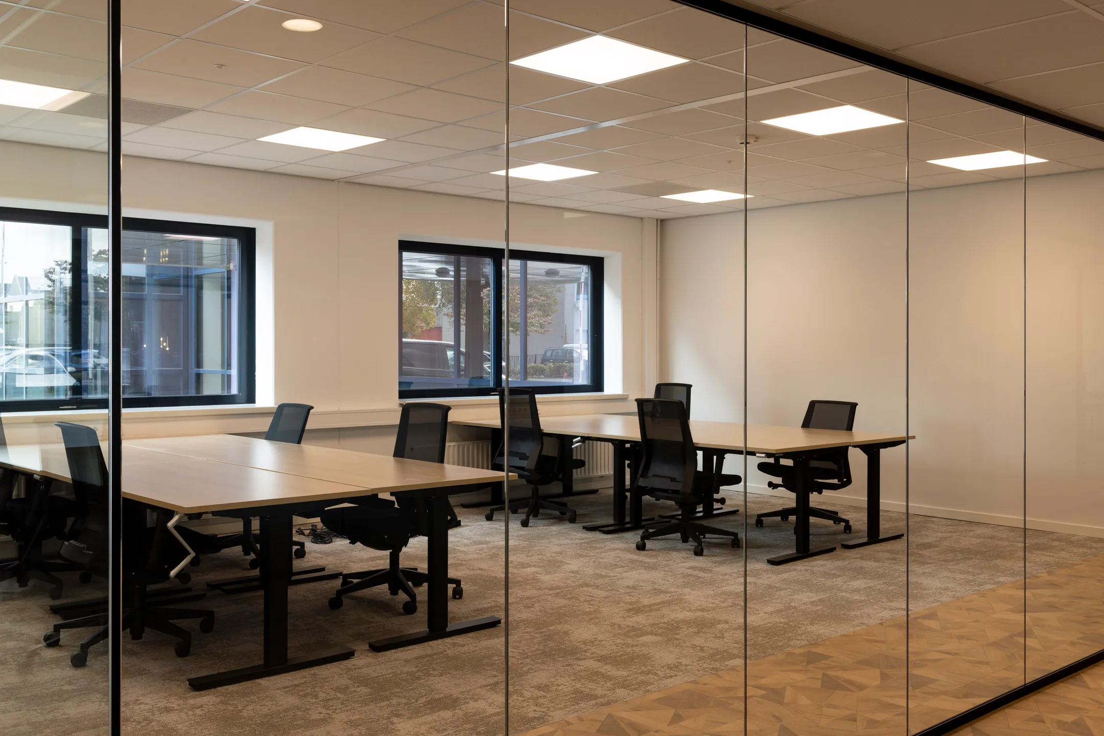 Een moderne vergaderruimte met glazen wanden, voorzien van zes bureaus en bureaustoelen.