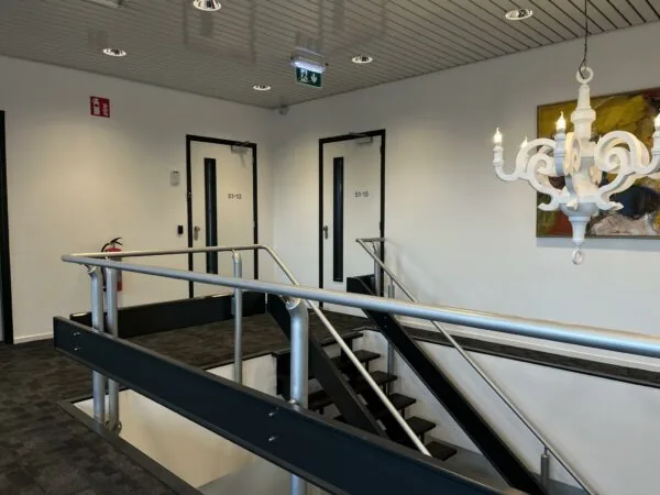 Trapopgang met metalen leuningen in een kantoorpand aan de Wattbaan, met zicht op deuren, kunstwerk en een witte decoratieve lamp.