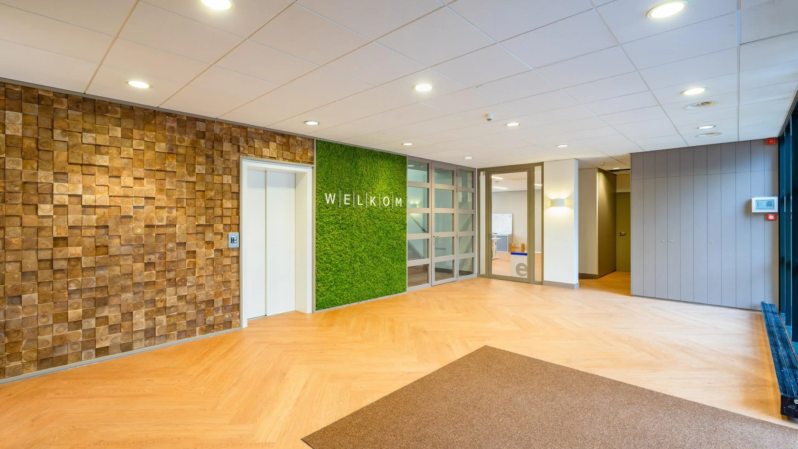 Ingang van een modern gebouw met houten vloer, groene moswand met "Welkom"-tekst, en houten decoratieve muurpanelen.