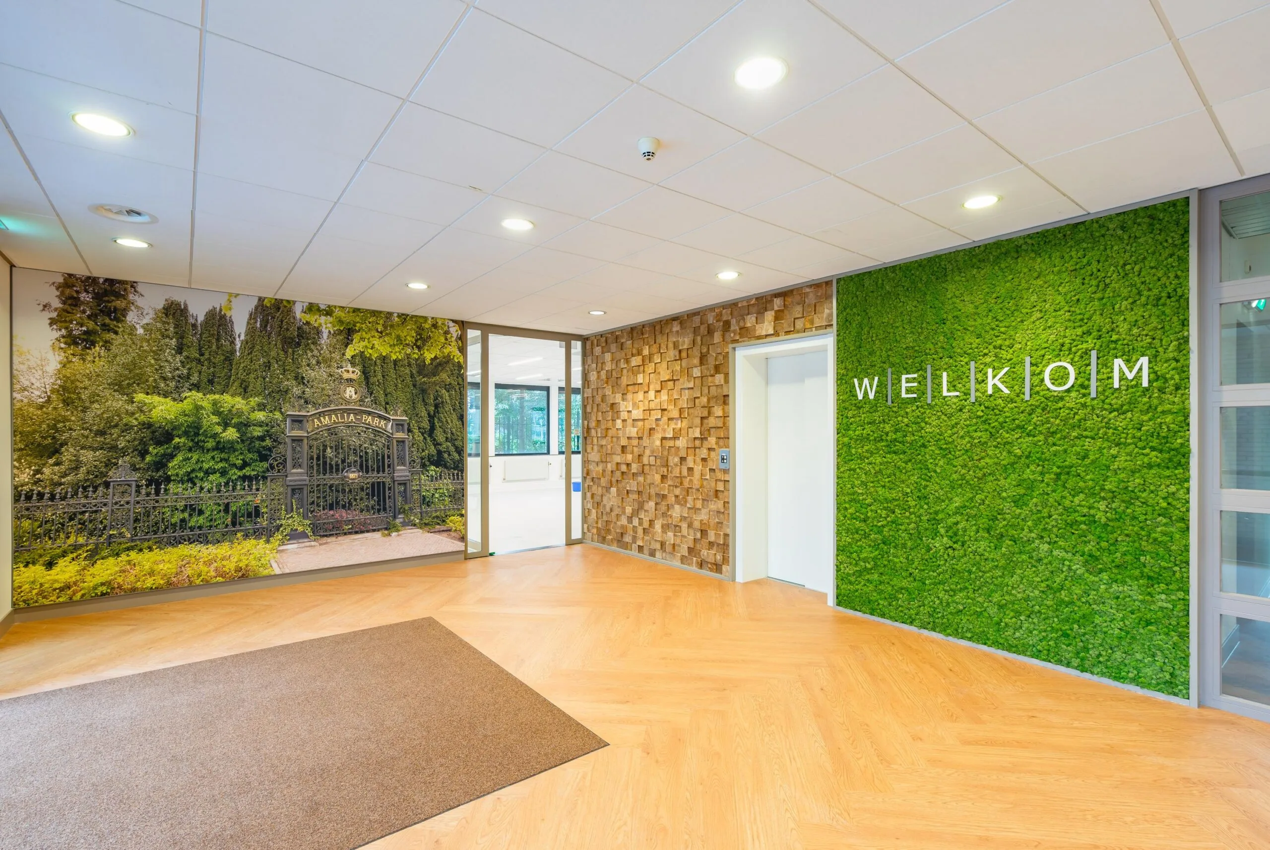 In een modern ingerichte ruimte hangt een grote fotomuur met de afbeelding van een parkpoort en een groene muur met de tekst "WELKOM".