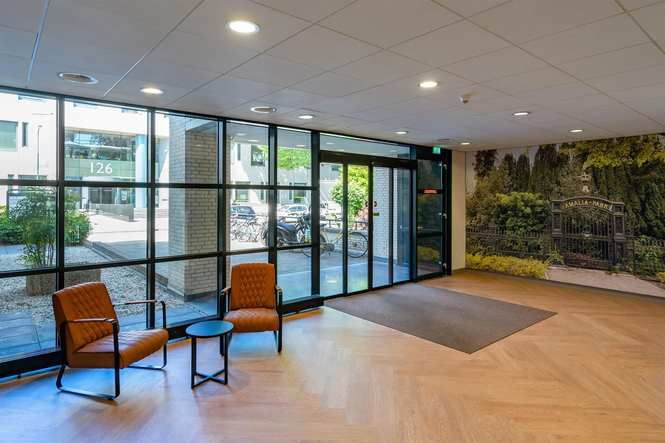 Entree van een gebouw met moderne stoelen, een glazen wand en een afbeelding van Amaliapark op de muur.