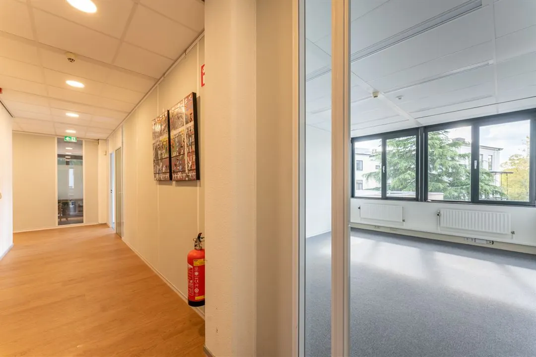 Een lichte gang met houten vloer, kunst aan de muur en een brandblusser, grenzend aan een kamer met grote ramen.