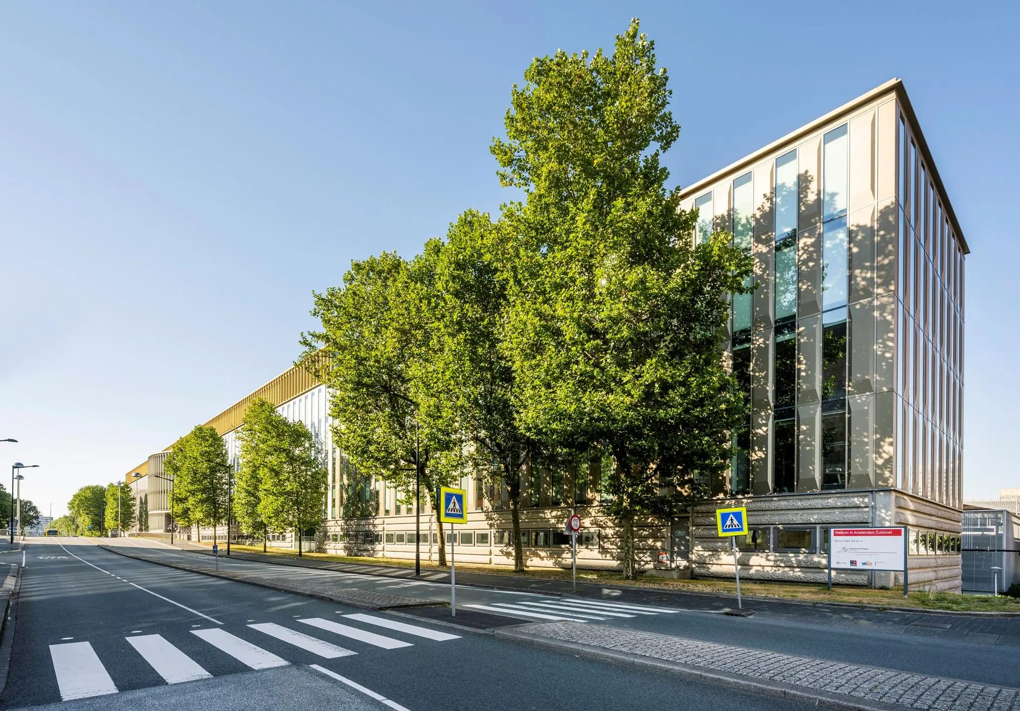 Modern kantoorgebouw aan de Hoekenrode in Amsterdam met groene bomen langs een rustige straat met zebrapaden.