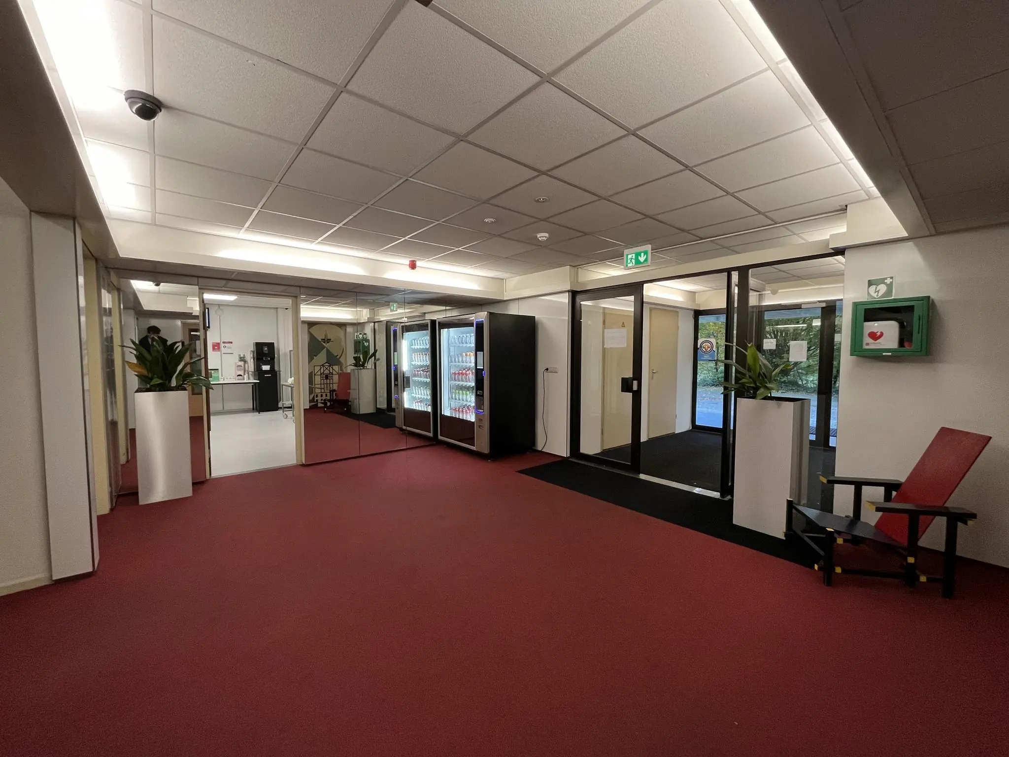Entreehal van een kantoorgebouw aan de Industrieweg met frisdrankautomaten, plantenbakken, een defibrillator en een rood-zwarte designstoel.