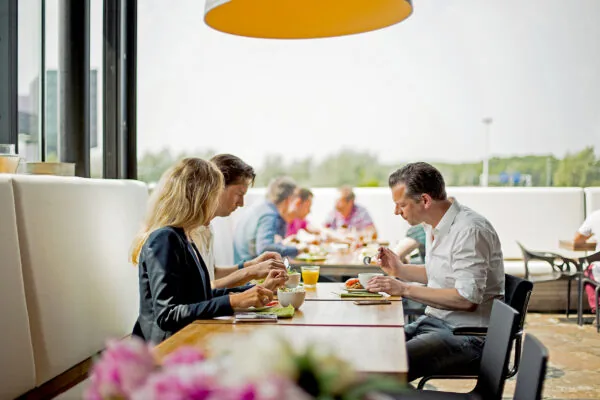 Mensen zitten samen te lunchen aan een lange tafel in een modern bedrijfsrestaurant aan de Orteliuslaan.