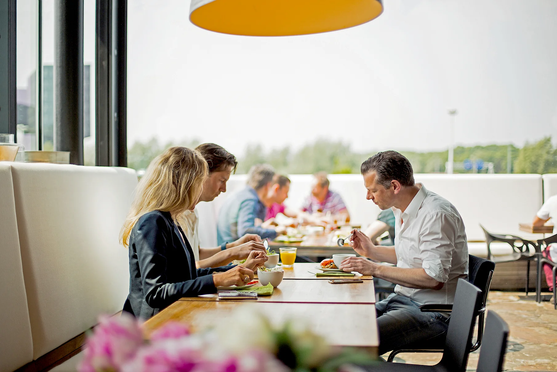 Mensen zitten samen te lunchen aan een lange tafel in een modern bedrijfsrestaurant aan de Orteliuslaan.