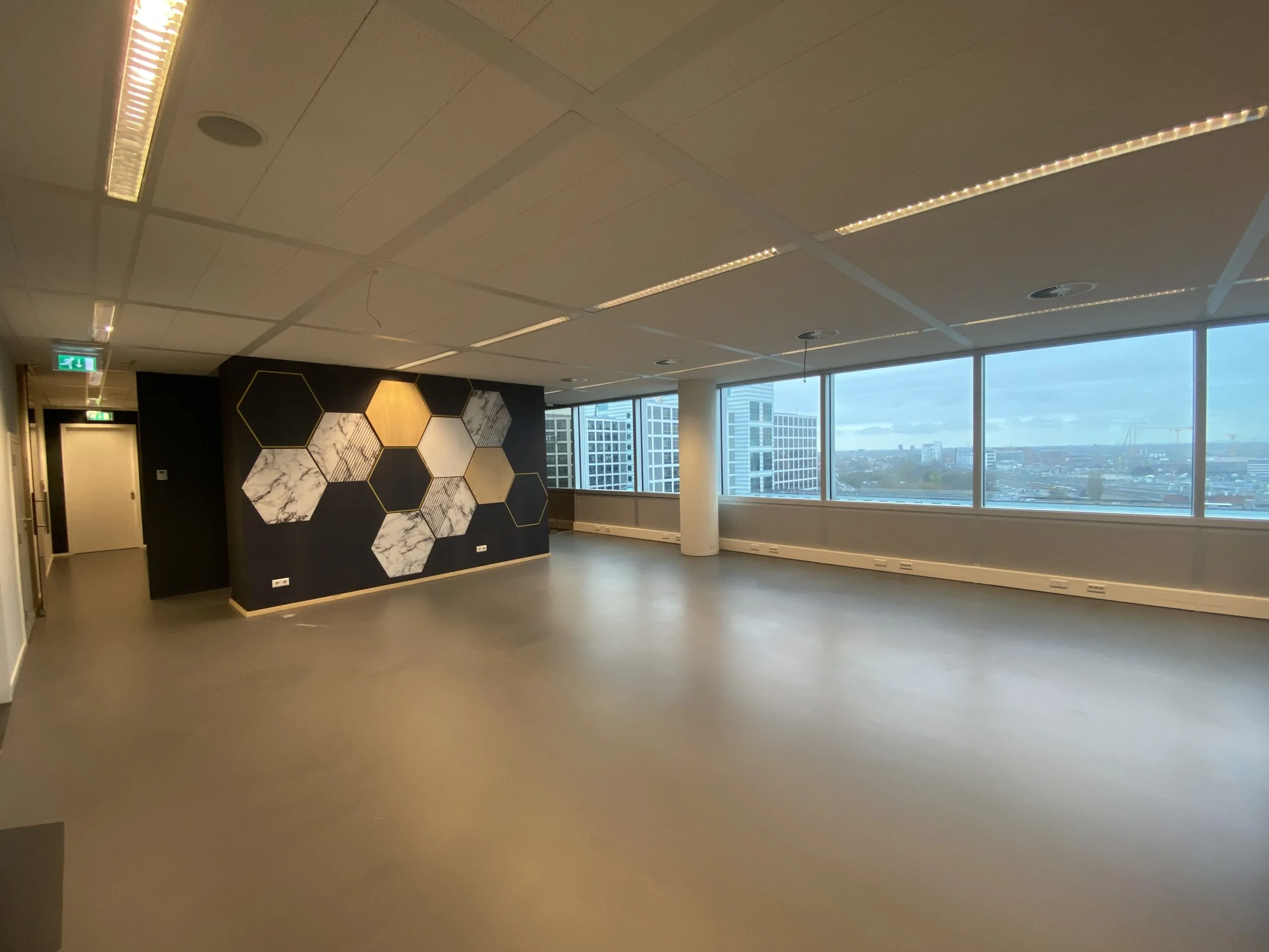 Lege, moderne kantoorruimte met grote ramen en een muurdecoratie van zeshoekige panelen in marmer, hout en zwart.