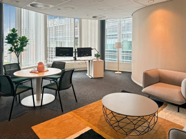 Een modern kantoor met een ronde tafel, stoelen, bureaus met monitoren, en een bank, omringd door grote ramen met uitzicht op de stad.
