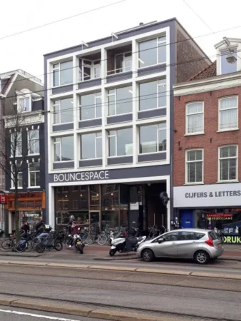 Een moderne vier verdiepingen tellend gebouw met de naam "BounceSpace" op de begane grond, omringd door fietsen en geparkeerde auto's.