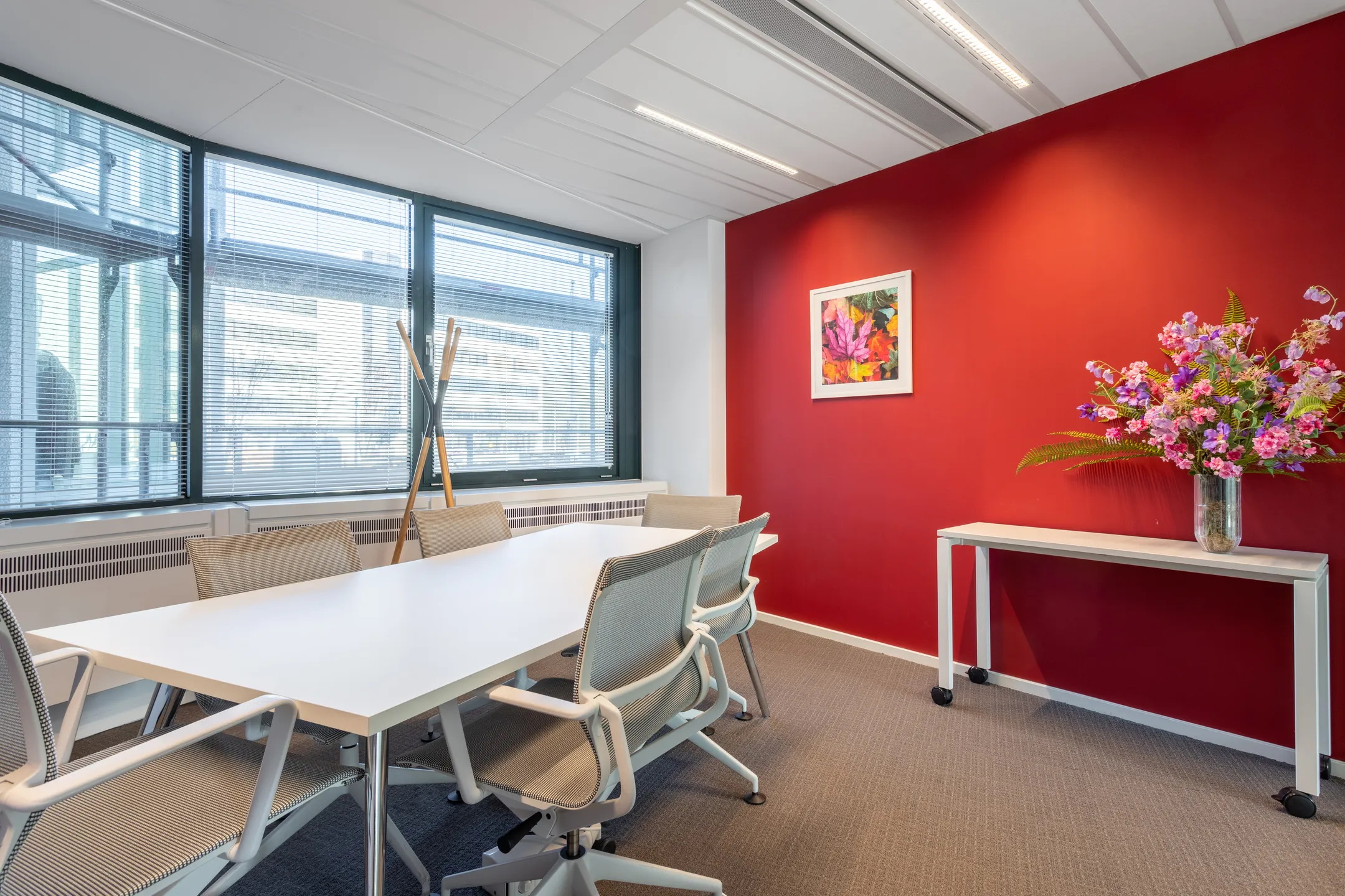 Een moderne vergaderruimte met een rode muur, een witte tafel en stoelen, bloemstuk op een bijzettafel en kleurrijk schilderij.