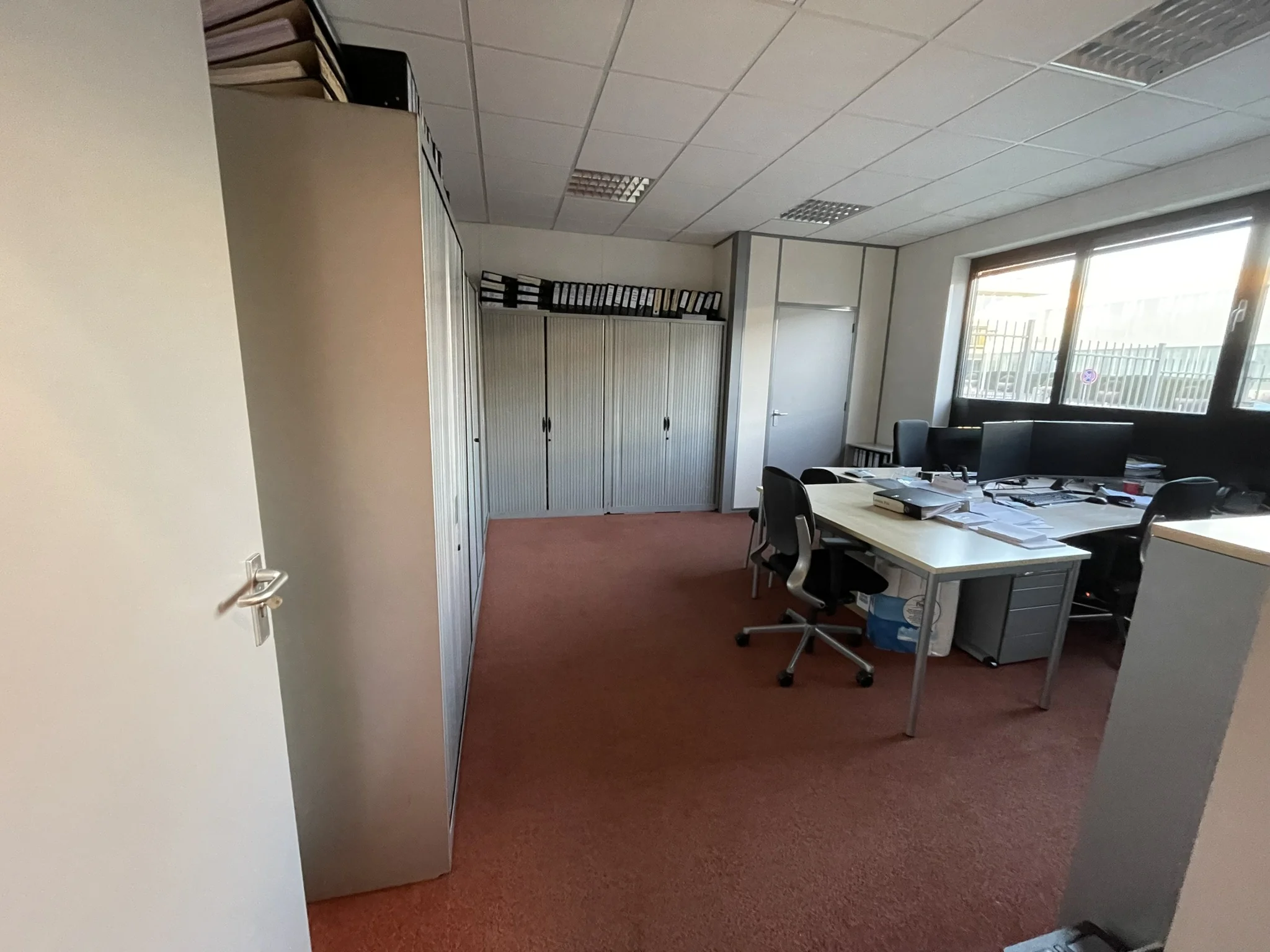 Kantoorruimte met bureaus, kantoormeubilair, archiefkasten en computers aan een roodtapijtvloer.