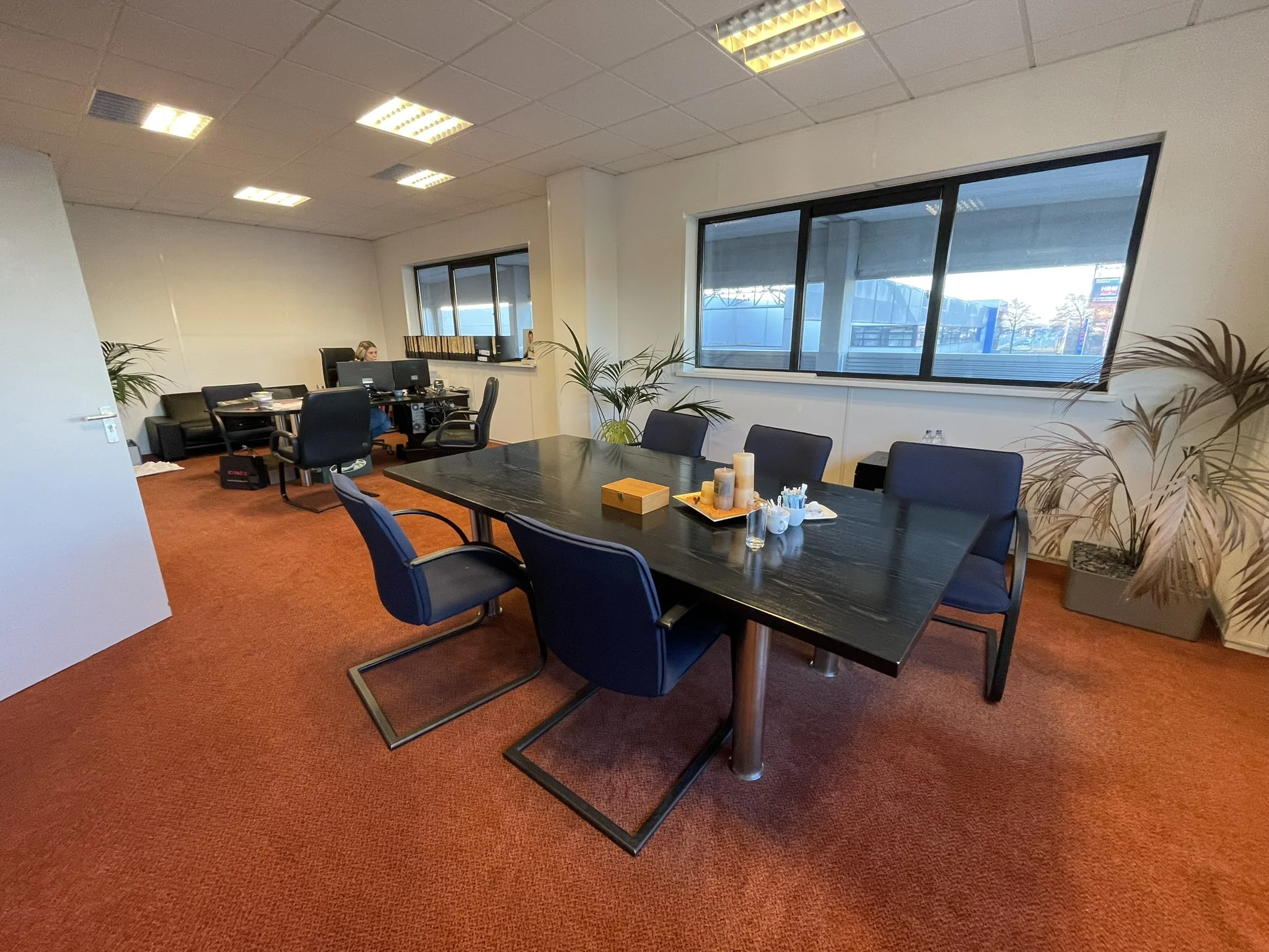 Moderne kantoorruimte met een vergadertafel, bureaus, stoelen en grote ramen met uitzicht naar buiten.