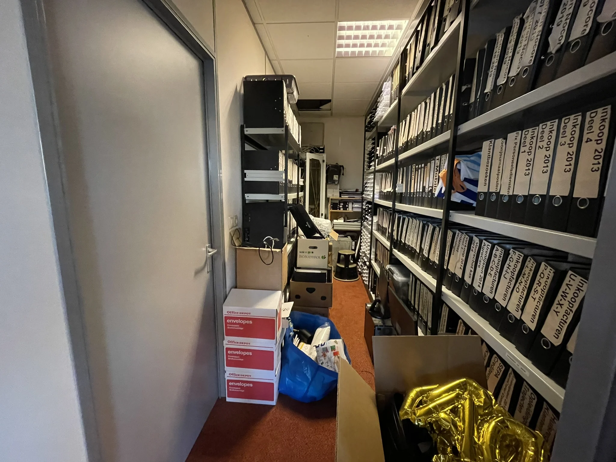 Volgepakte archiefruimte met rijen ordners, dozen en losse spullen op de vloer.
