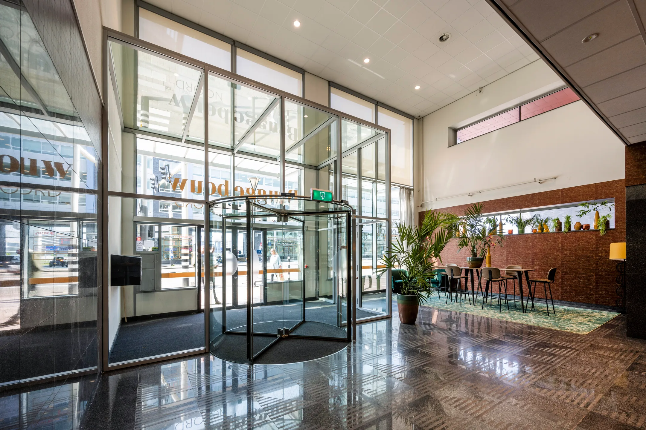 Ingang van een modern kantoorgebouw met een glazen draaideur en een zitgedeelte met planten.