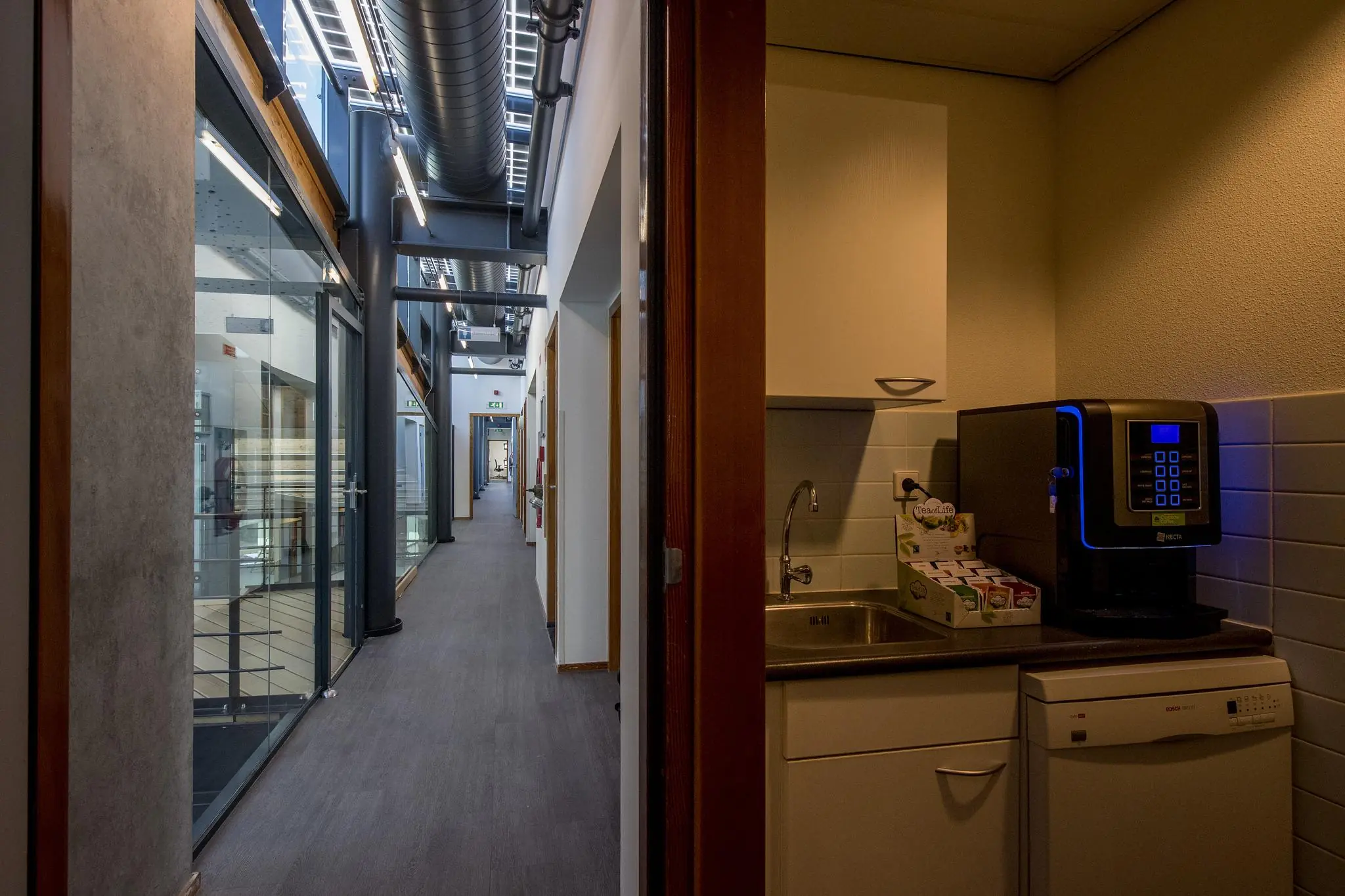 Gang in een modern kantoorpand aan de Fokkerstraat met glazen wanden en een pantry met koffiemachine.
