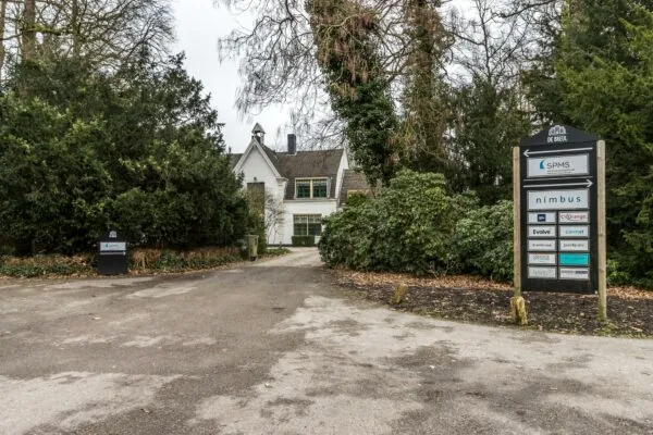 Oprijlaan naar een wit kantoorpand aan de Driebergseweg met omringend groen en een bord met bedrijfsnamen.
