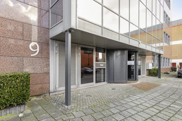 Ingang van een modern kantoorpand aan de Curieweg 9 met een draaiende toegangsdeur en spiegelende gevel.