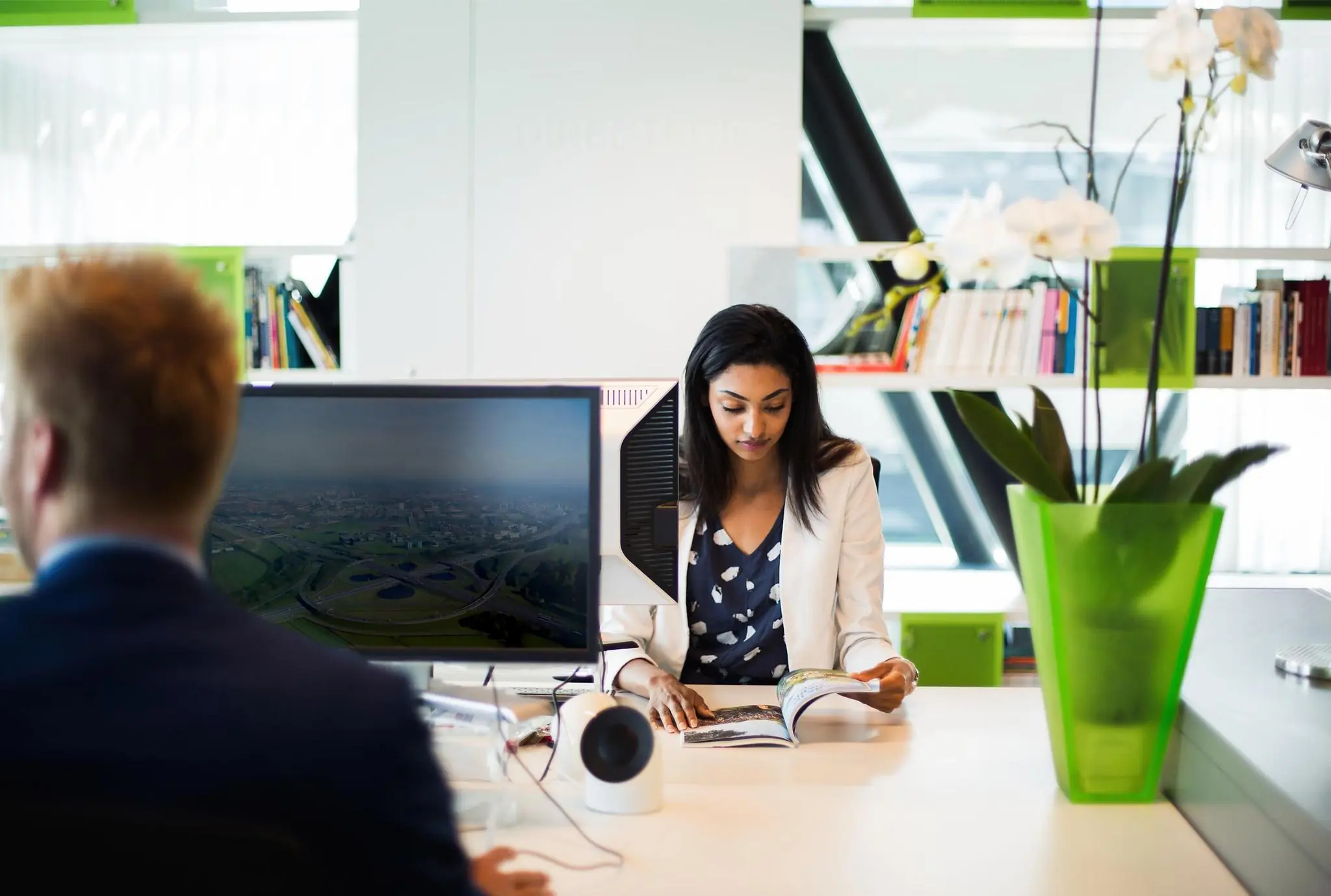 Twee mensen werken op een modern kantoor aan de Orteliuslaan, met een vrouw die een tijdschrift leest en een man achter een computerscherm.