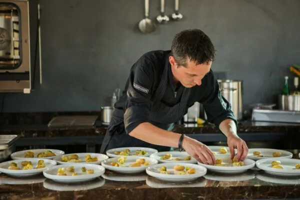 Chef-kok in zwarte kledij bereidt meerdere borden opmaakgerechten in een professionele keuken aan de Orteliuslaan.