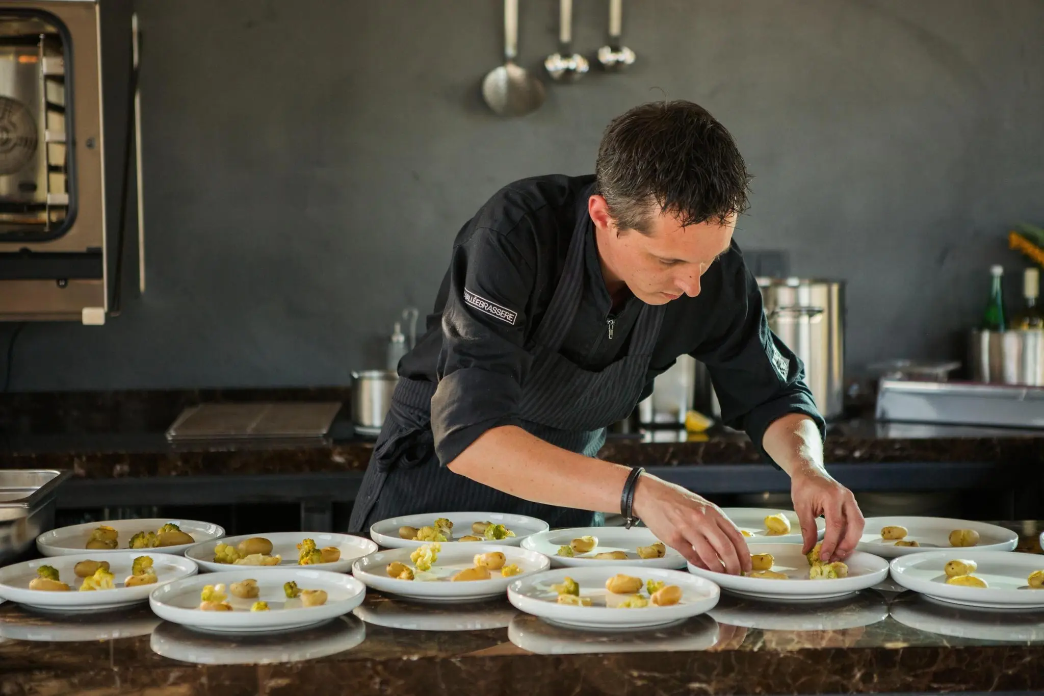 Chef-kok in zwarte kledij bereidt meerdere borden opmaakgerechten in een professionele keuken aan de Orteliuslaan.