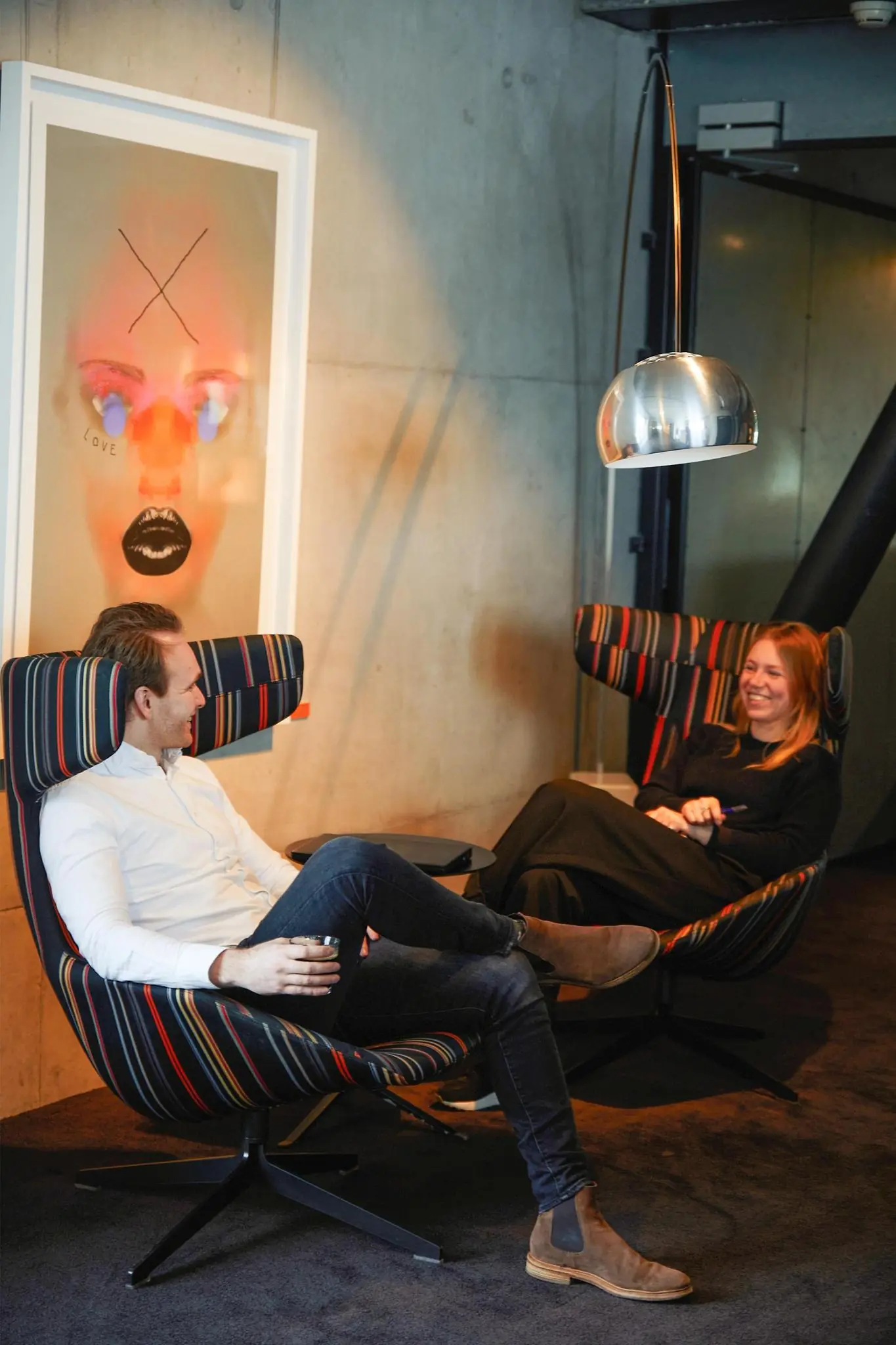 Twee mensen zitten ontspannen in designstoelen bij Orteliuslaan, in gesprek onder een moderne lamp en naast kunst aan de muur.