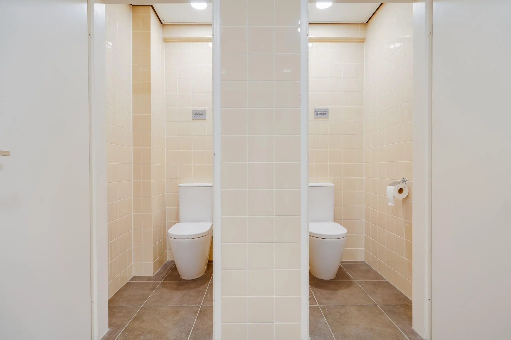 Twee moderne, naast elkaar gelegen toiletcabines met beige wandtegels en witte deuren in een gebouw aan de Zonnebaan.