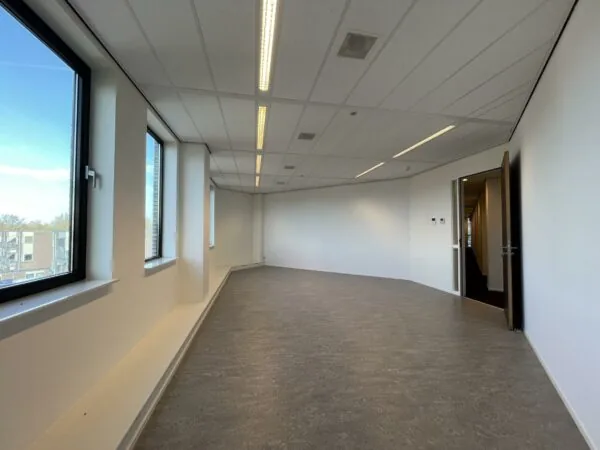 Leeg kantoorruimte met grote ramen, witte muren en een open deur naar een gang in het gebouw Ratelaar.