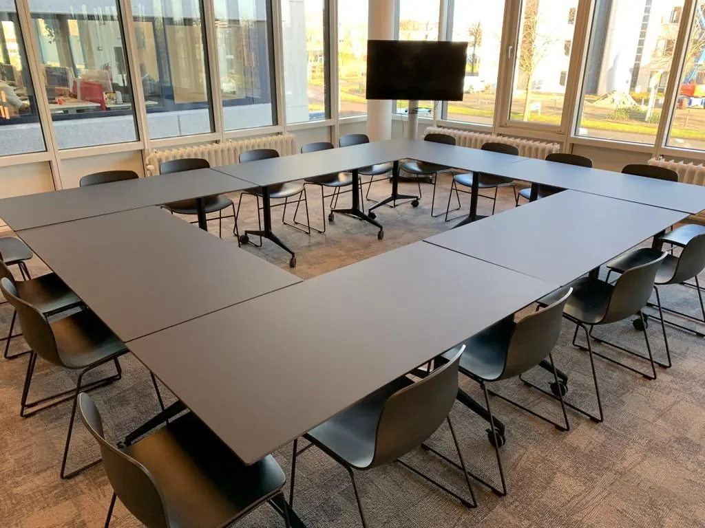 Vergaderruimte in Rijnzathe met een U-vormige opstelling van zwarte tafels en stoelen, omringd door grote ramen en voorzien van een presentatiescherm.