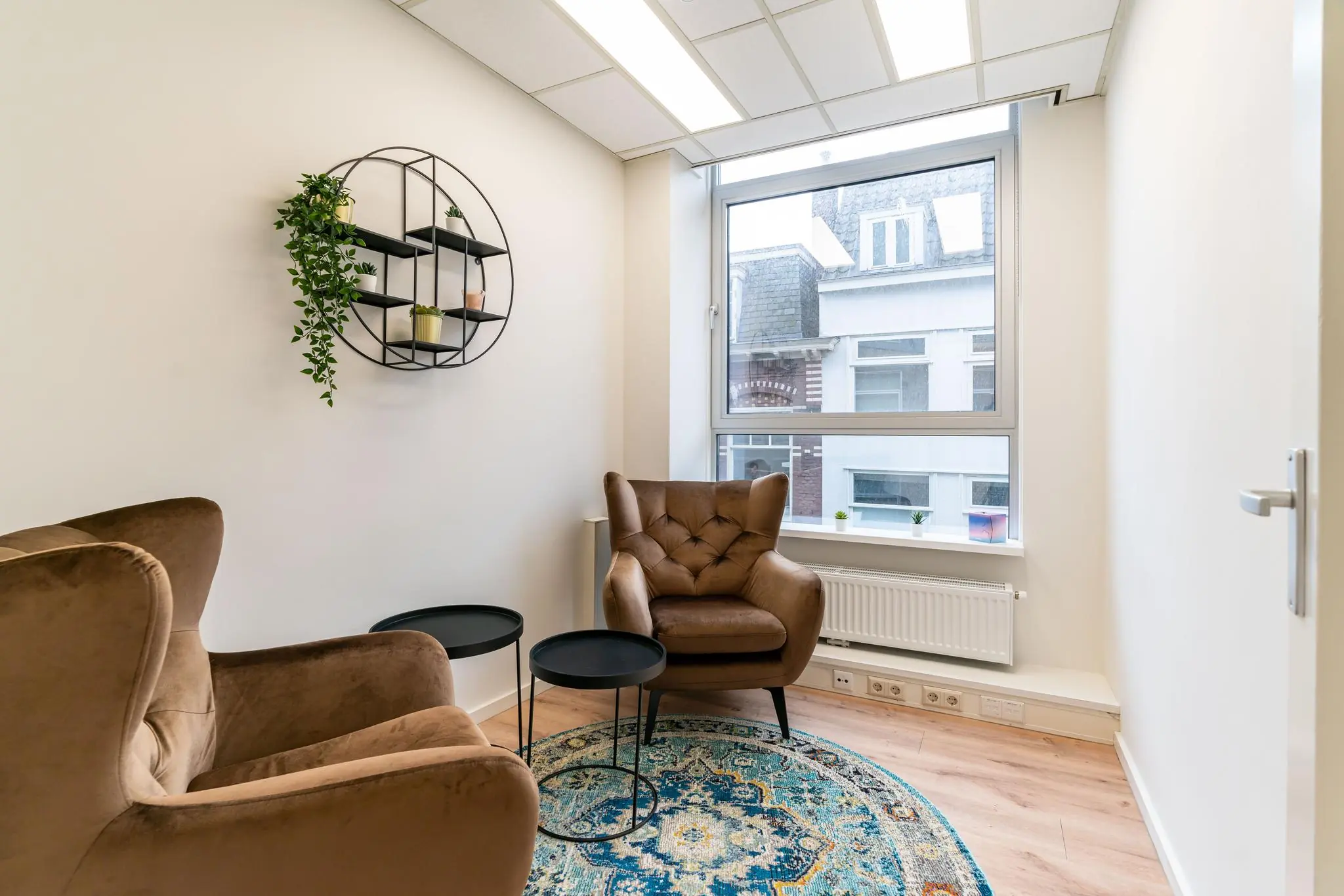 Kleine, modern ingerichte spreekkamer met twee bruine fauteuils, een rond vloerkleed en een groot raam met uitzicht op panden aan het Plein.