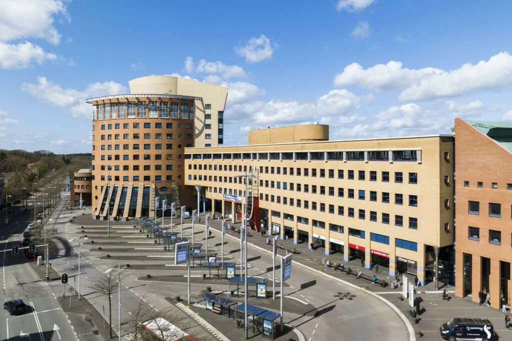 Modern kantoorgebouw aan het Stationsplein met bushaltes en voetgangers op een zonnige dag.