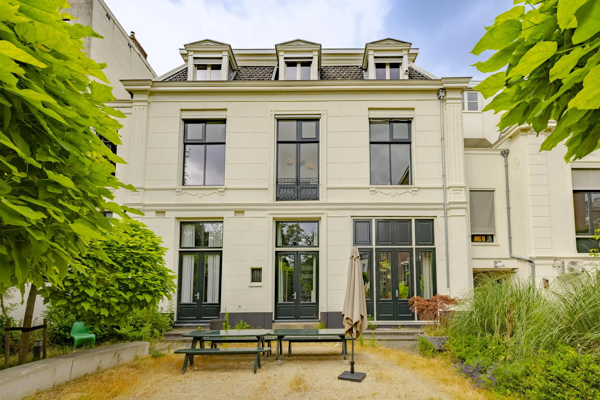 Achtergevel van een statig wit herenhuis aan de Plantage Middenlaan met hoge ramen, een tuin met picknicktafels en omliggend groen.