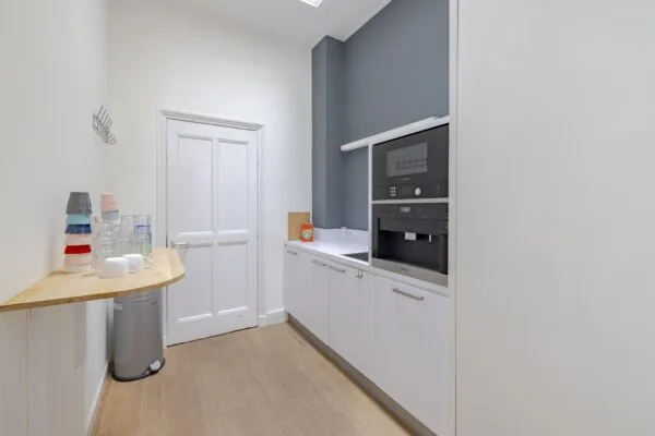 Kleine pantry met ingebouwde koffiemachine, aanrechtblad en stapels bekers op een smalle houten tafel.