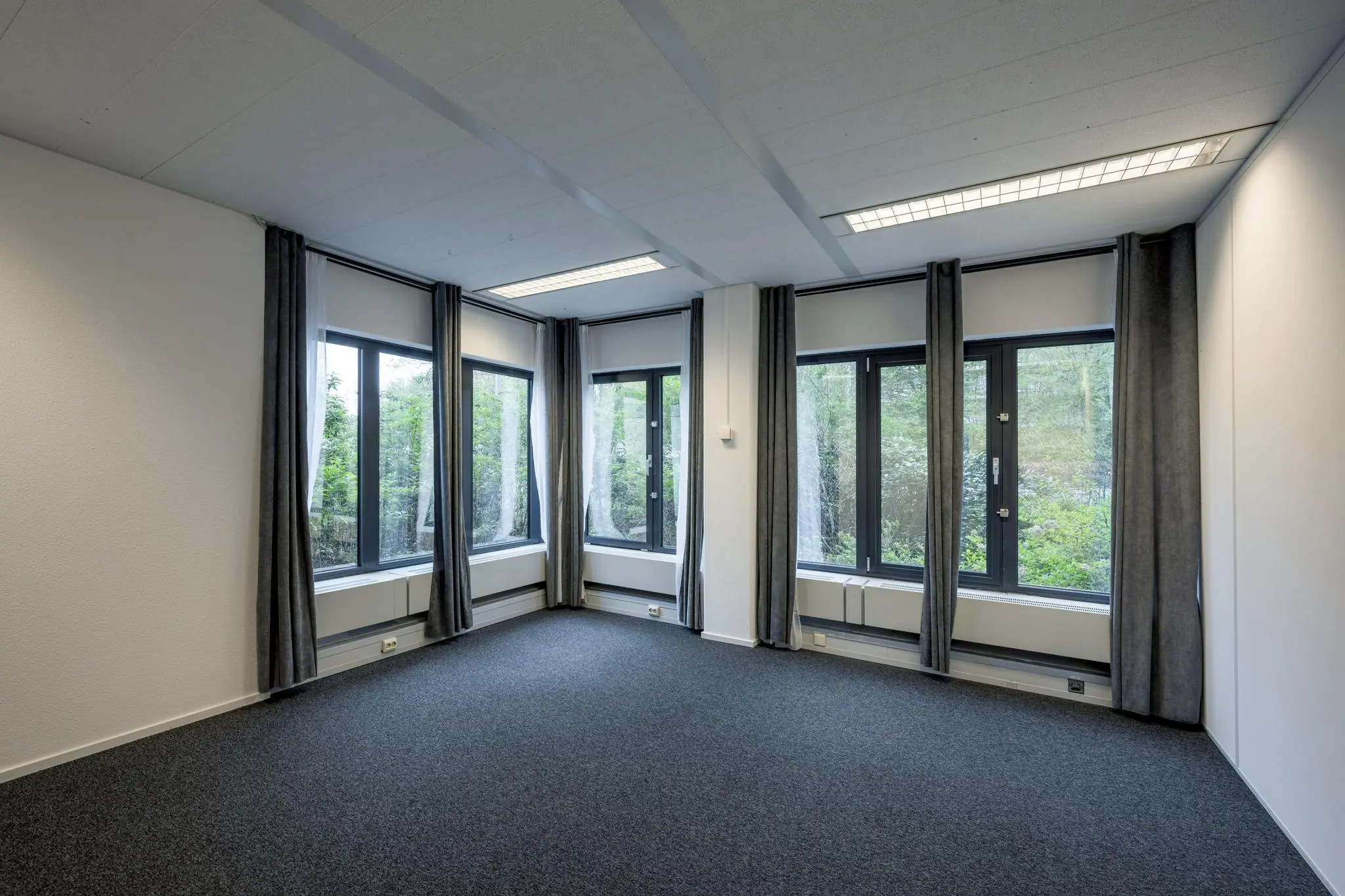 Licht en leeg kantoor met grote ramen, grijze gordijnen en uitzicht op groen.