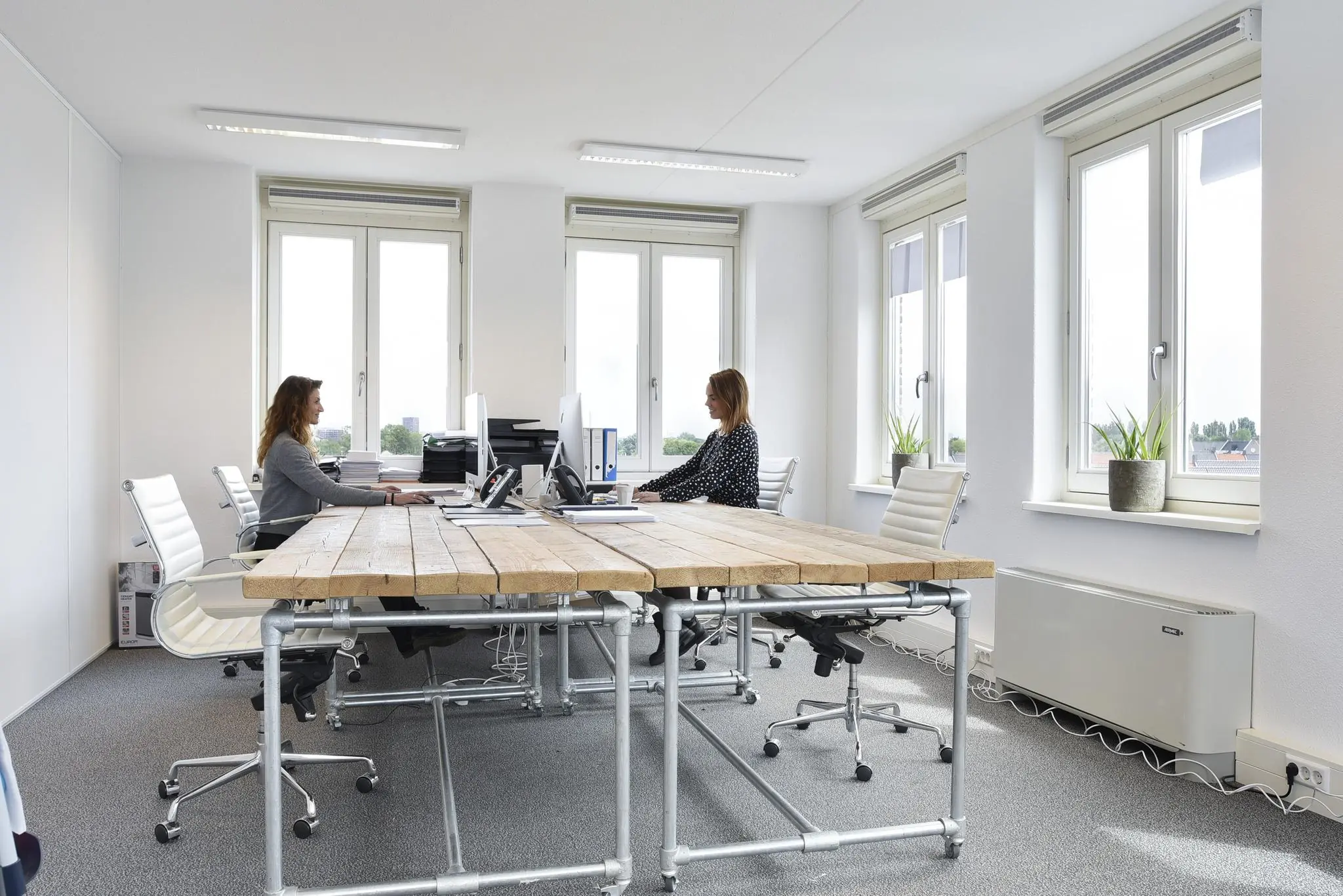 Twee vrouwen werken tegenover elkaar aan een houten vergadertafel in een licht, modern kantoor in Middenburcht.