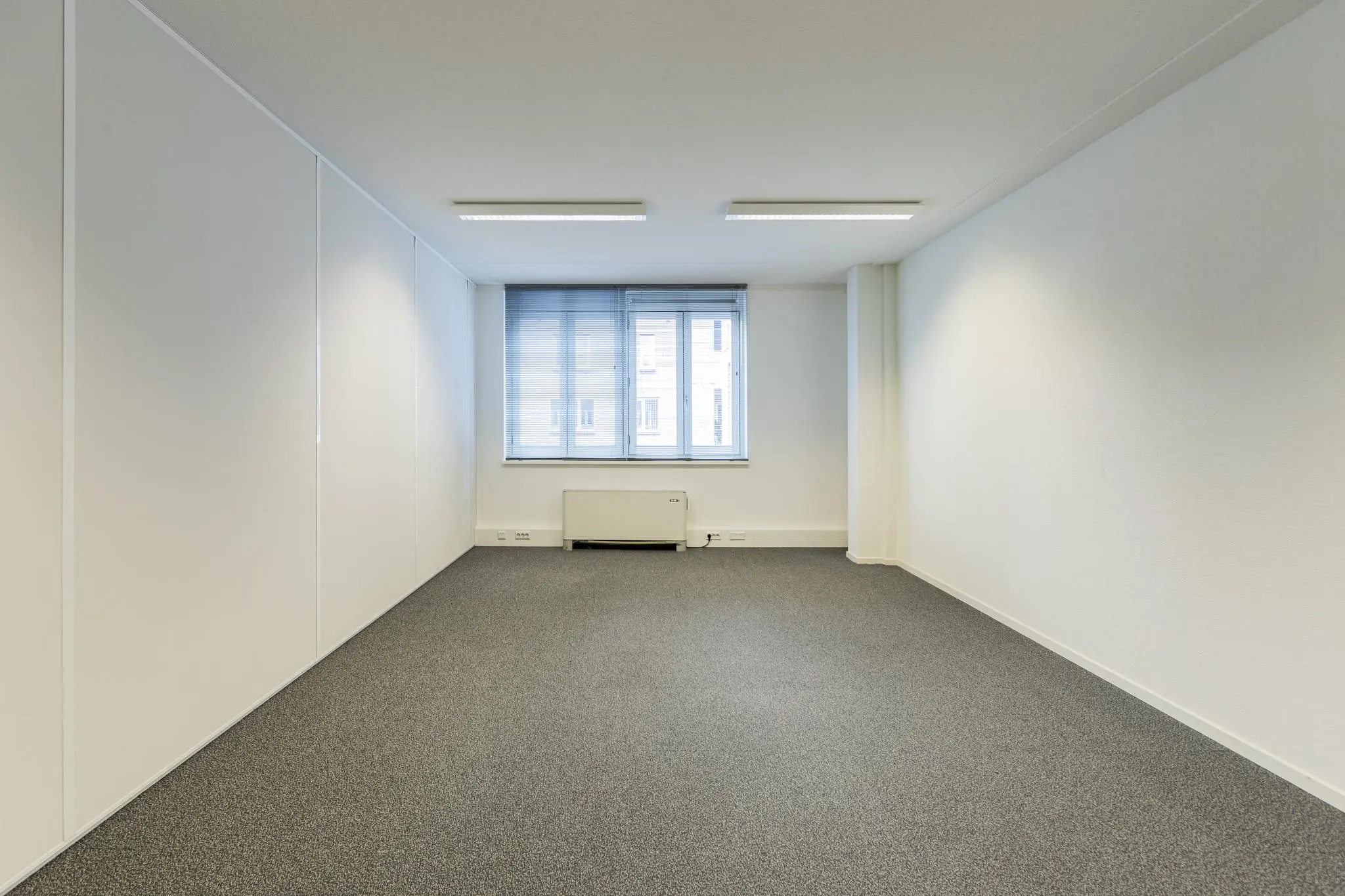 Lege, lichte kantoorruimte in het gebouw Middenburcht met grijs tapijt en een raam met jaloezieën.