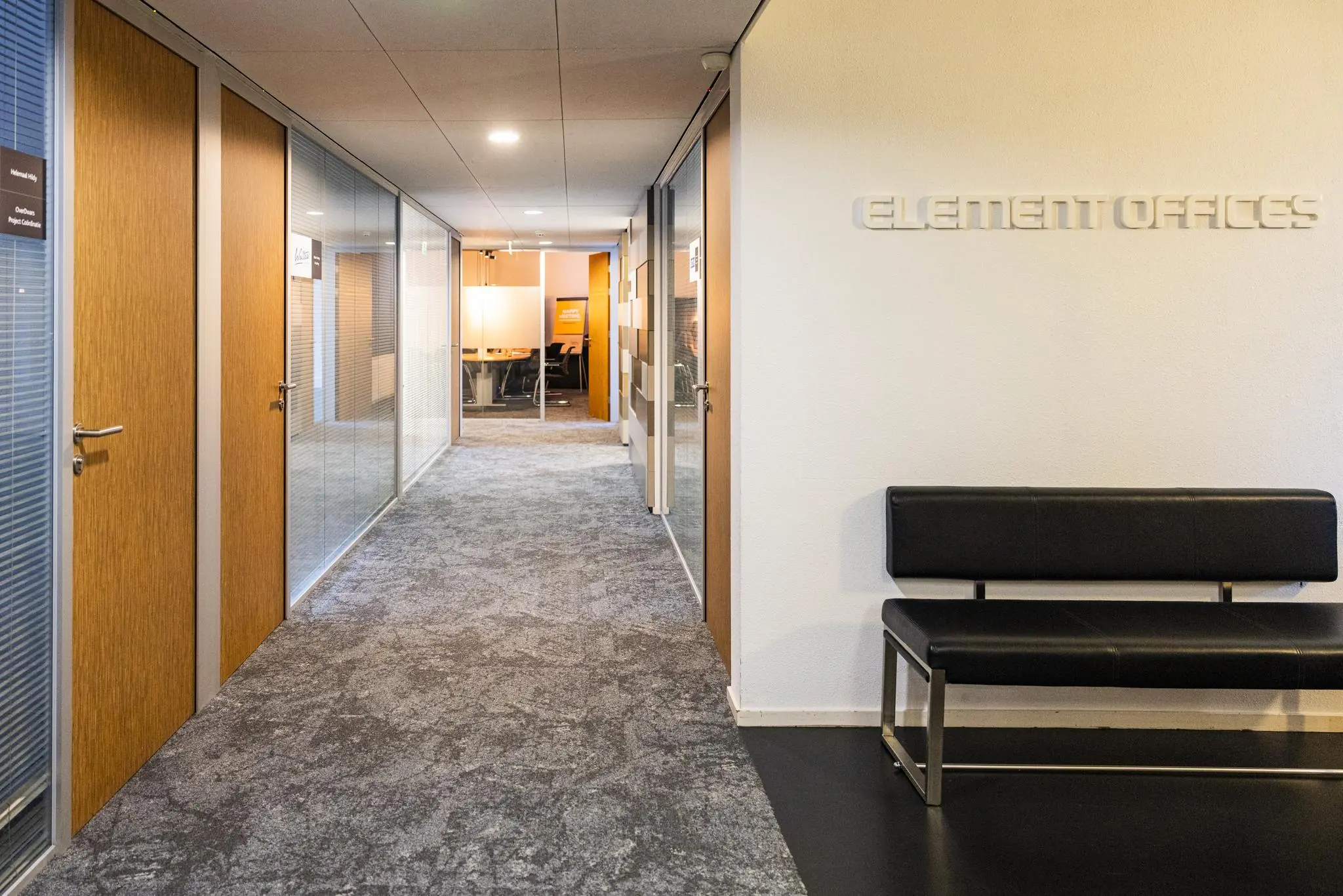 Gang in het kantoorgebouw Element Offices met glazen wanden, houten deuren en een zwarte wachtbank.