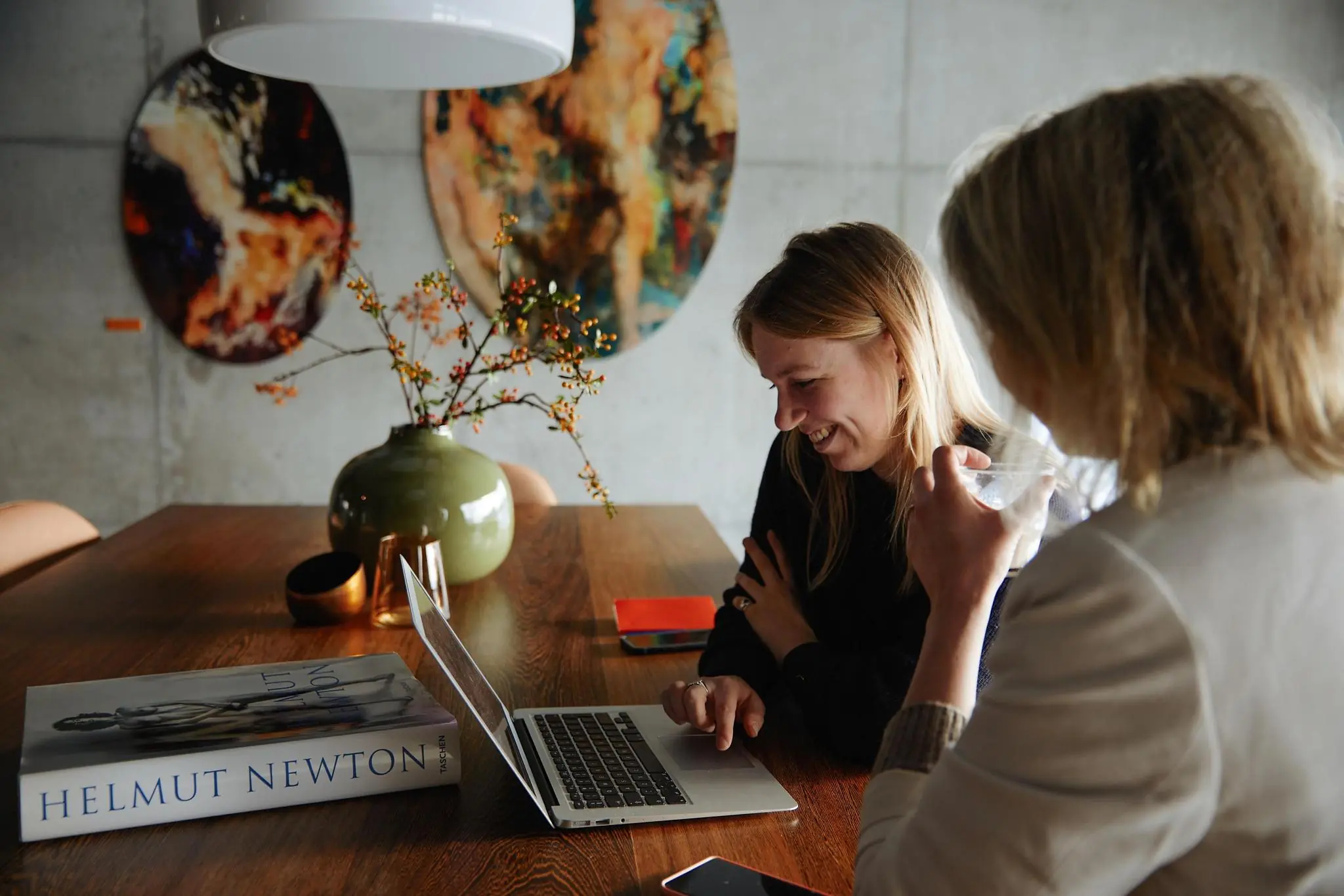 Twee vrouwen zitten lachend aan een tafel met een laptop in een modern interieur aan de Orteliuslaan.